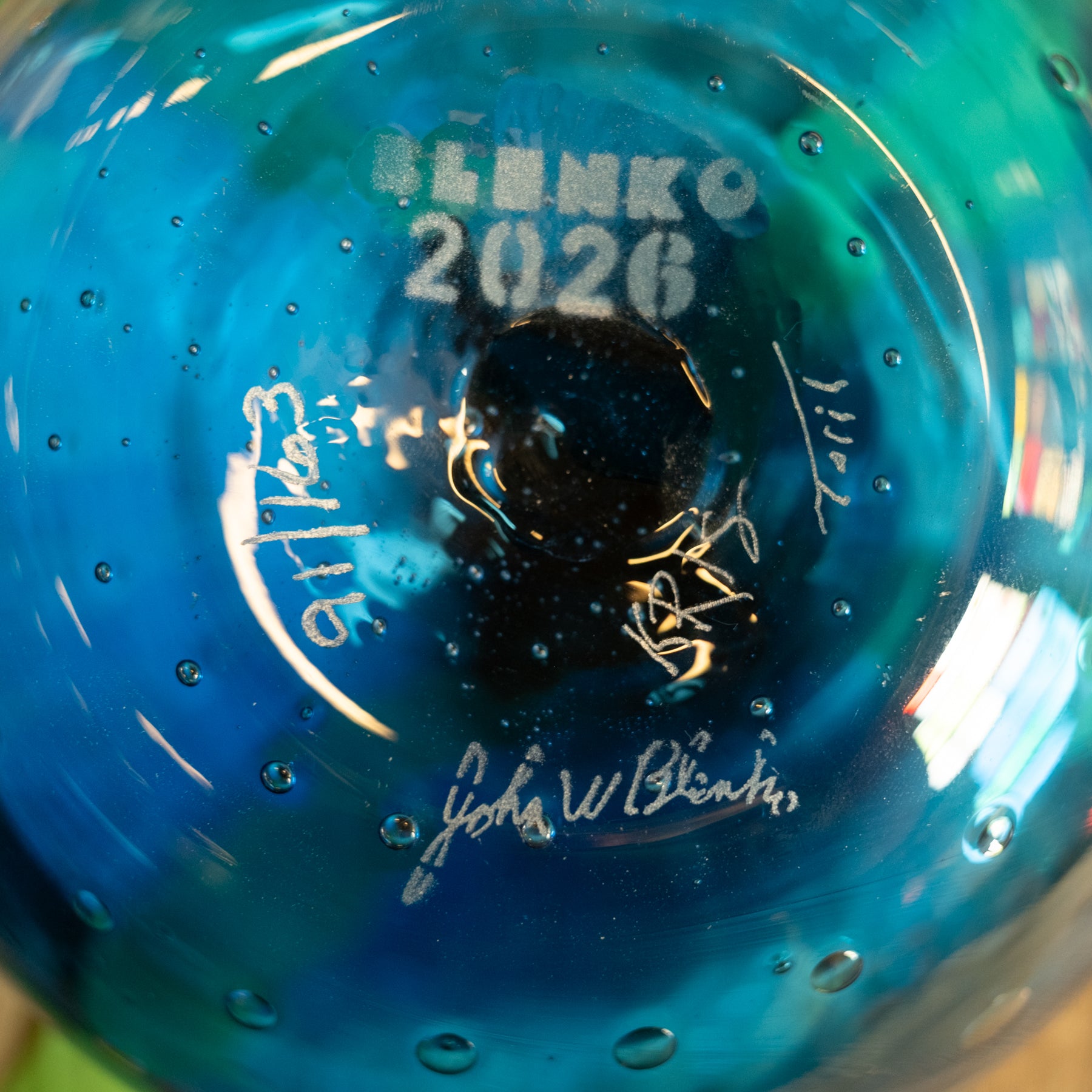 Close-up of the bottom of a blue glass bowl or vase with water droplets, featuring the text 'BENKO 2026' and several artist signatures in white and gold.