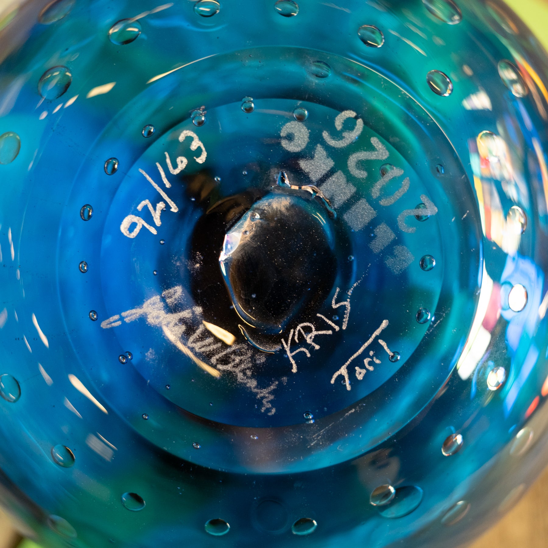 Close-up of a blue glass bowl or vase base with embossed circular patterns, handwritten white markings including dates and signatures, and a glossy reflective center.