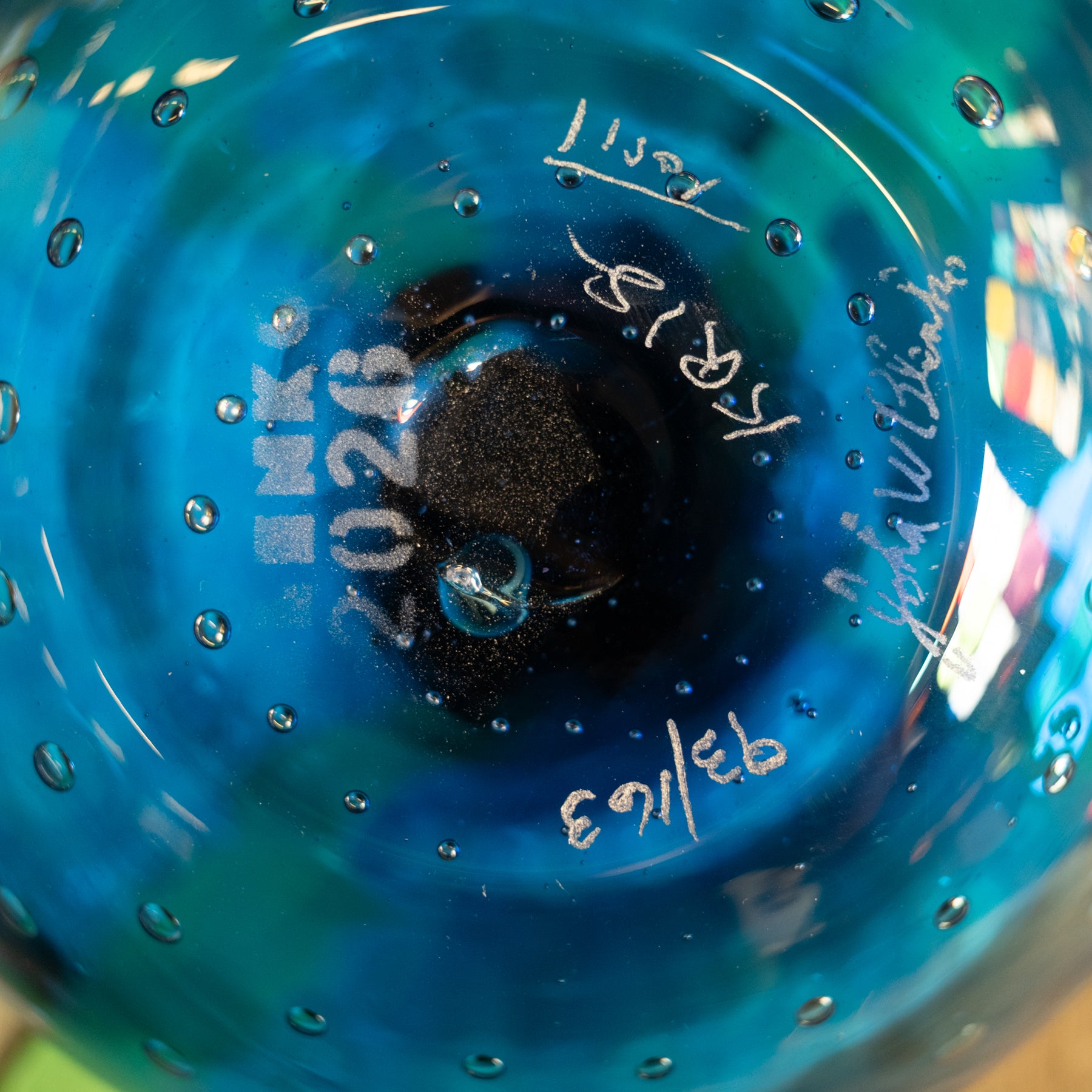 Close-up of the bottom of a blue glass bowl or vase with bubble details, showing white hand-written markings and a visible 'INK' brand stamp.