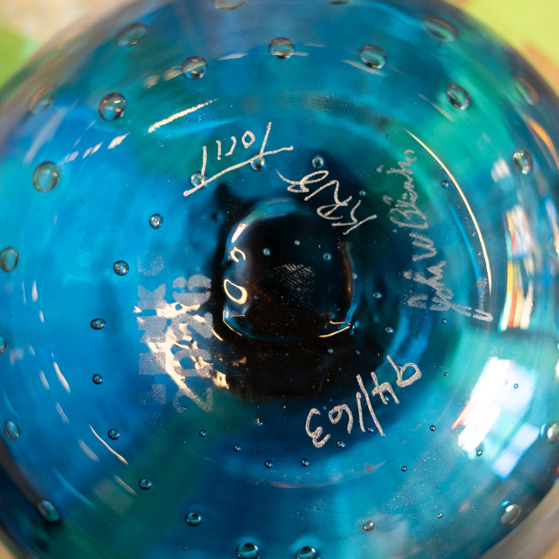 Close-up of the bottom of a blue glass bottle or vase with a bubbled texture and white handwritten markings.