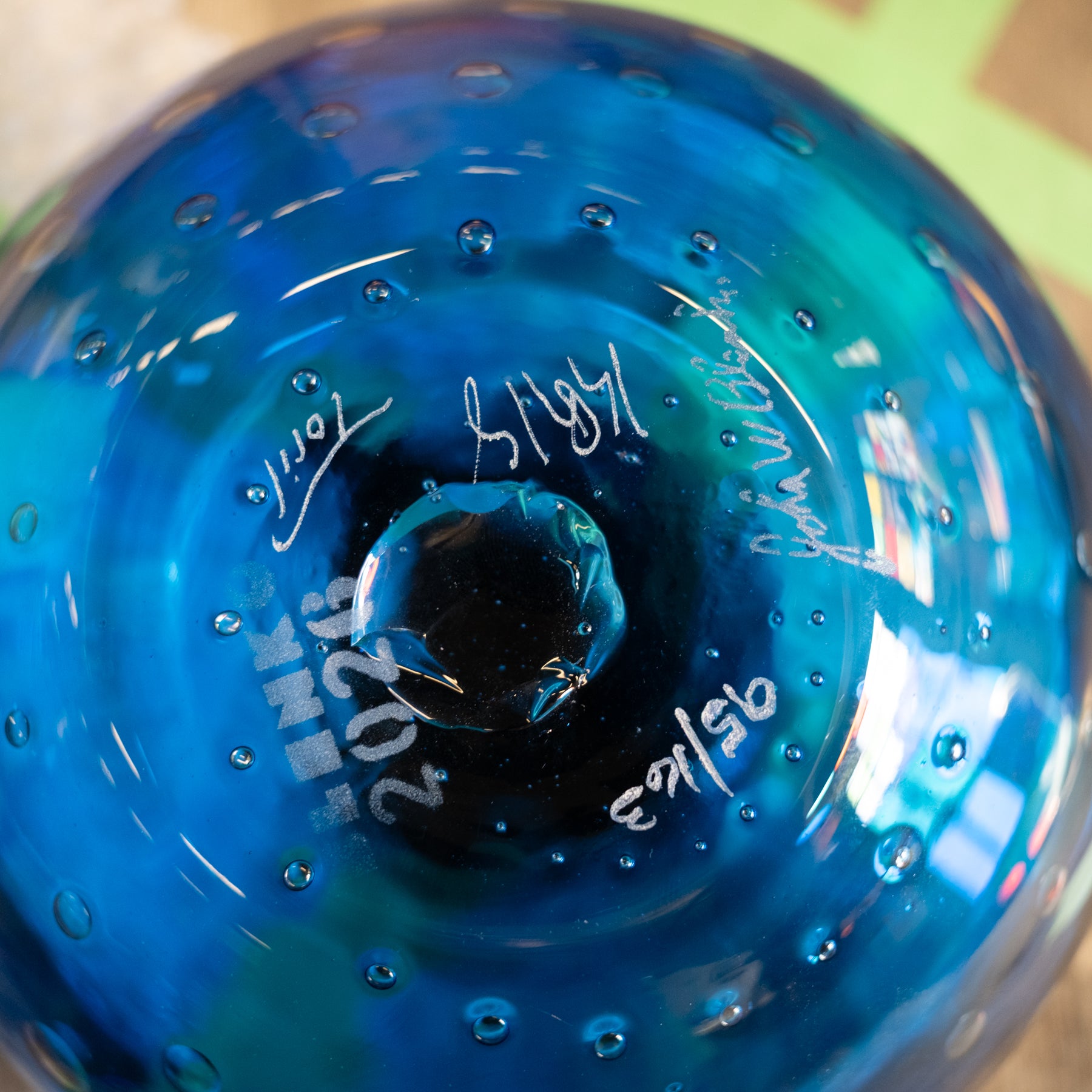 Close-up of the bottom of a blue glass vase or decorative glass object with a rounded bulbous shape, showing white handwritten markings and bubble details on a blurred background.