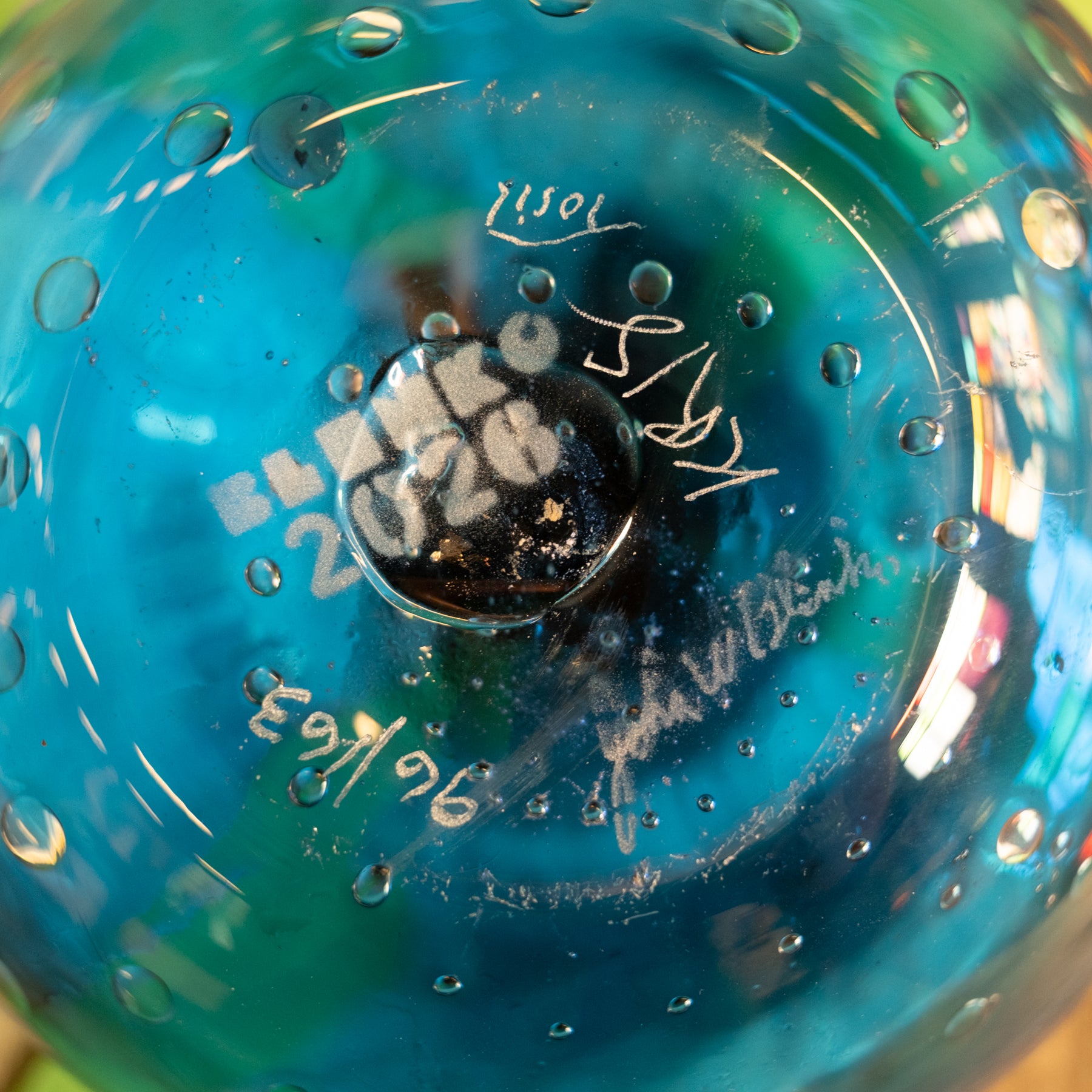 Close-up of the bottom of a transparent blue glass vessel with internal bubble detailing, featuring white printed text reading 'BENZO 2026' and several handwritten signatures.