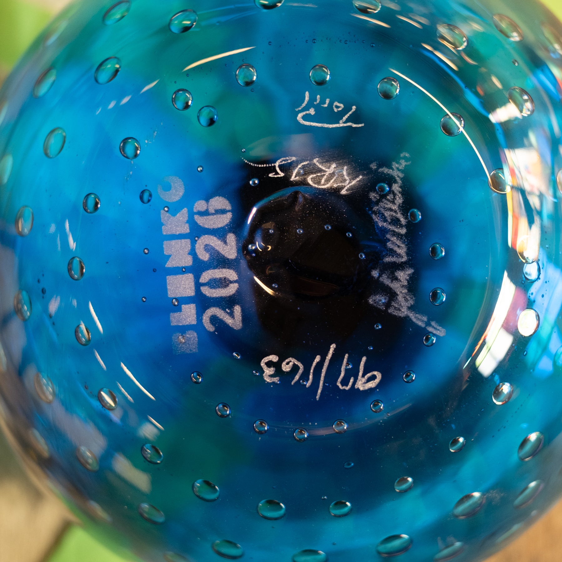 Close-up of the bottom of a translucent blue glass bowl with a textured bubble pattern, featuring white printed maker's marks and dates.