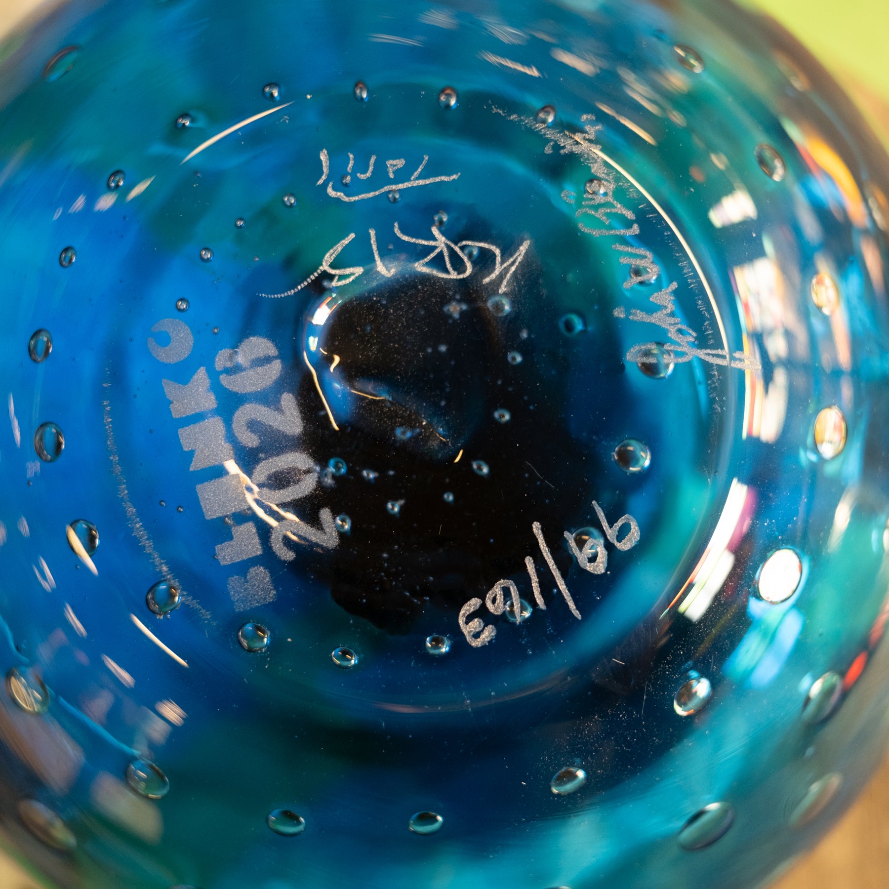 Close-up of the bottom of a blue glass bottle or vase with embossed text reading 'INKO 2026' and handwritten markings, showing a glossy transparent blue surface with small bubble details.