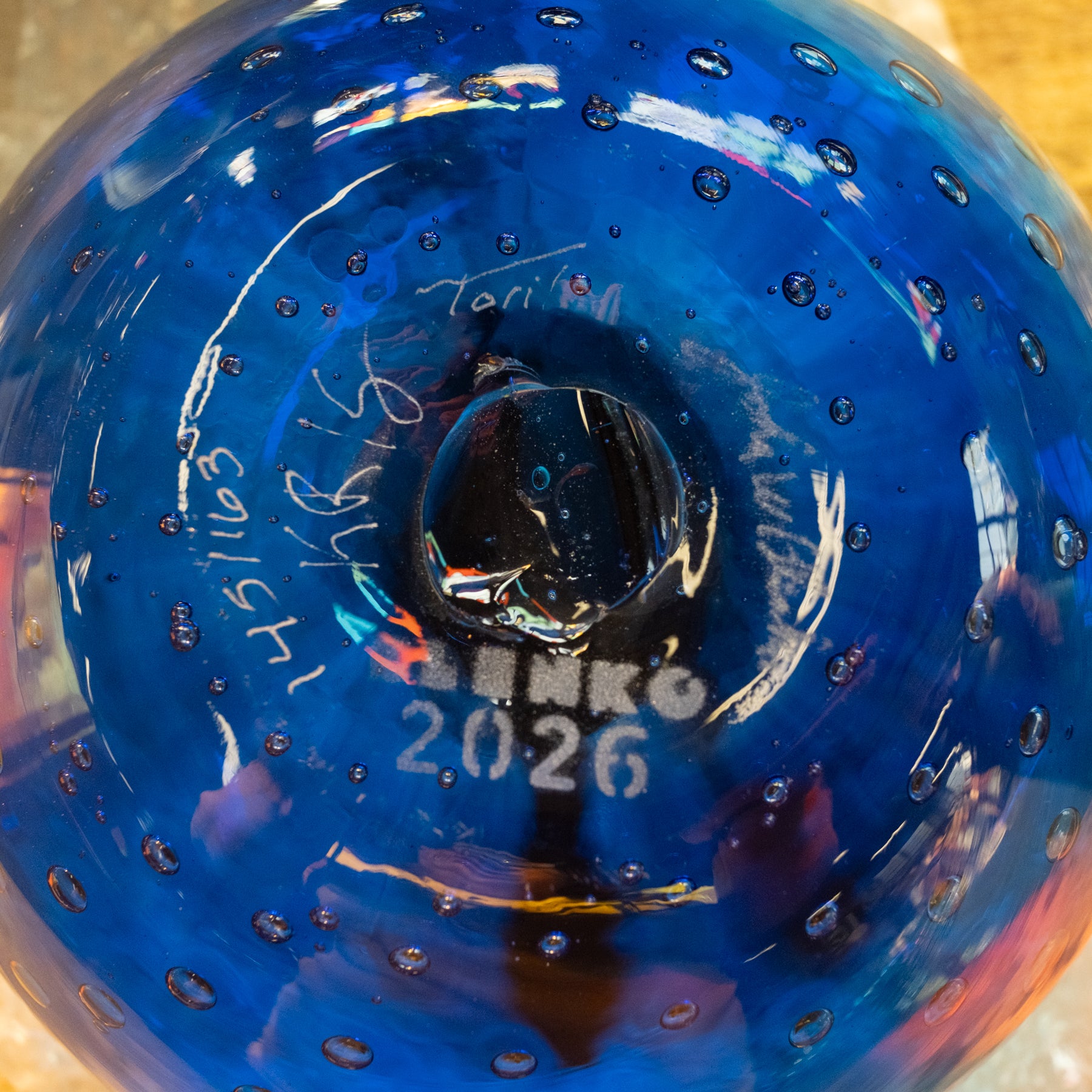 A close-up of a blue glass bottle or vase with a textured bubble pattern and white printed text reading 'Toni M' and '2026' on the bottom.