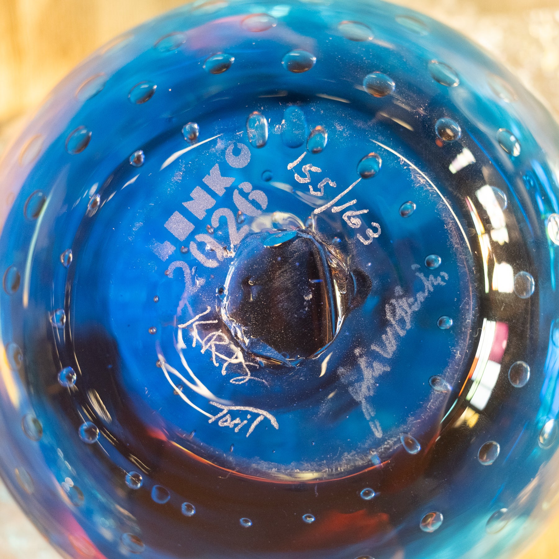 Close-up of the bottom of a blue glass bottle or vase with embossed white text and bubble details, photographed against a softly blurred warm beige background.