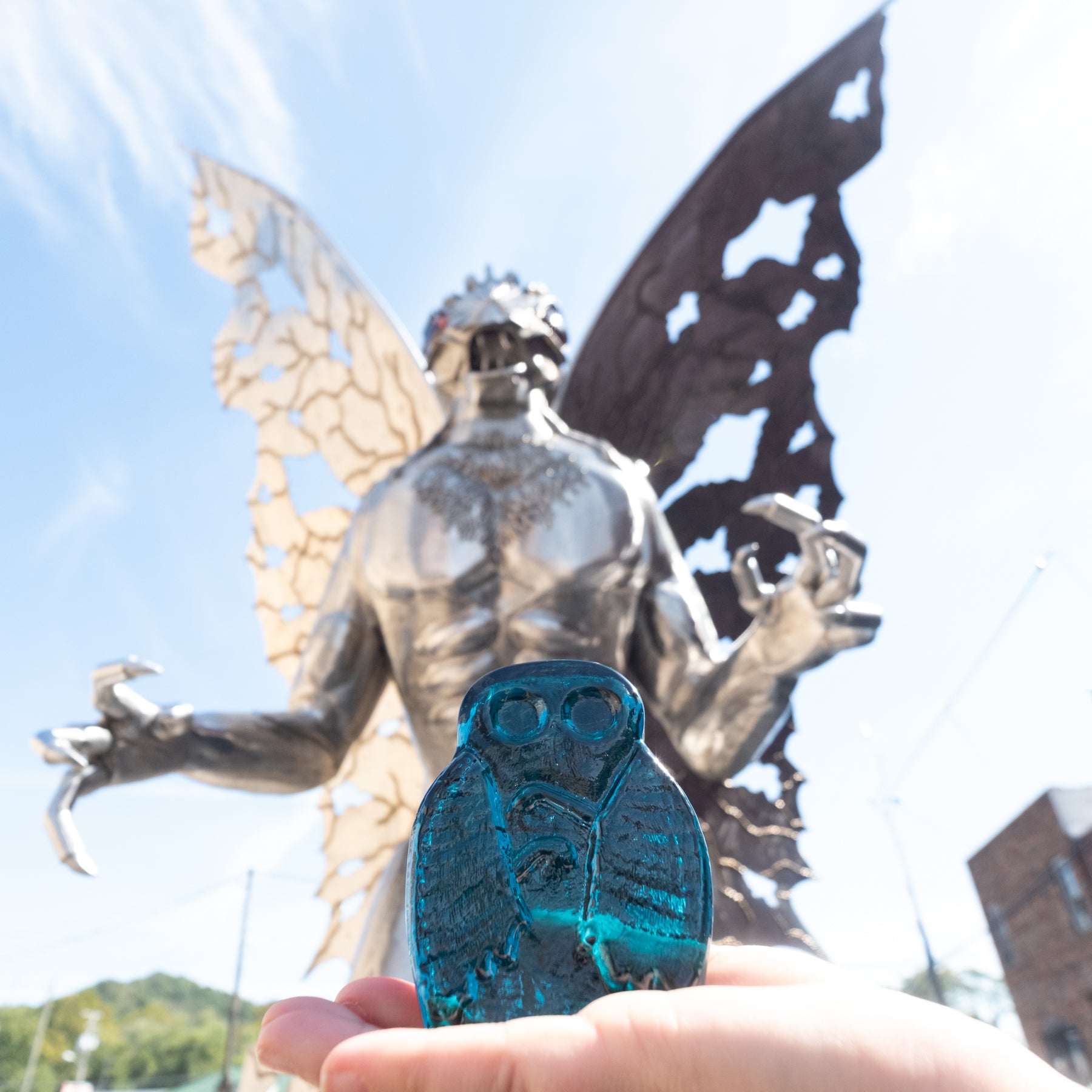 Person holding a blue object with an owl design in front of a large statue against a clear sky.