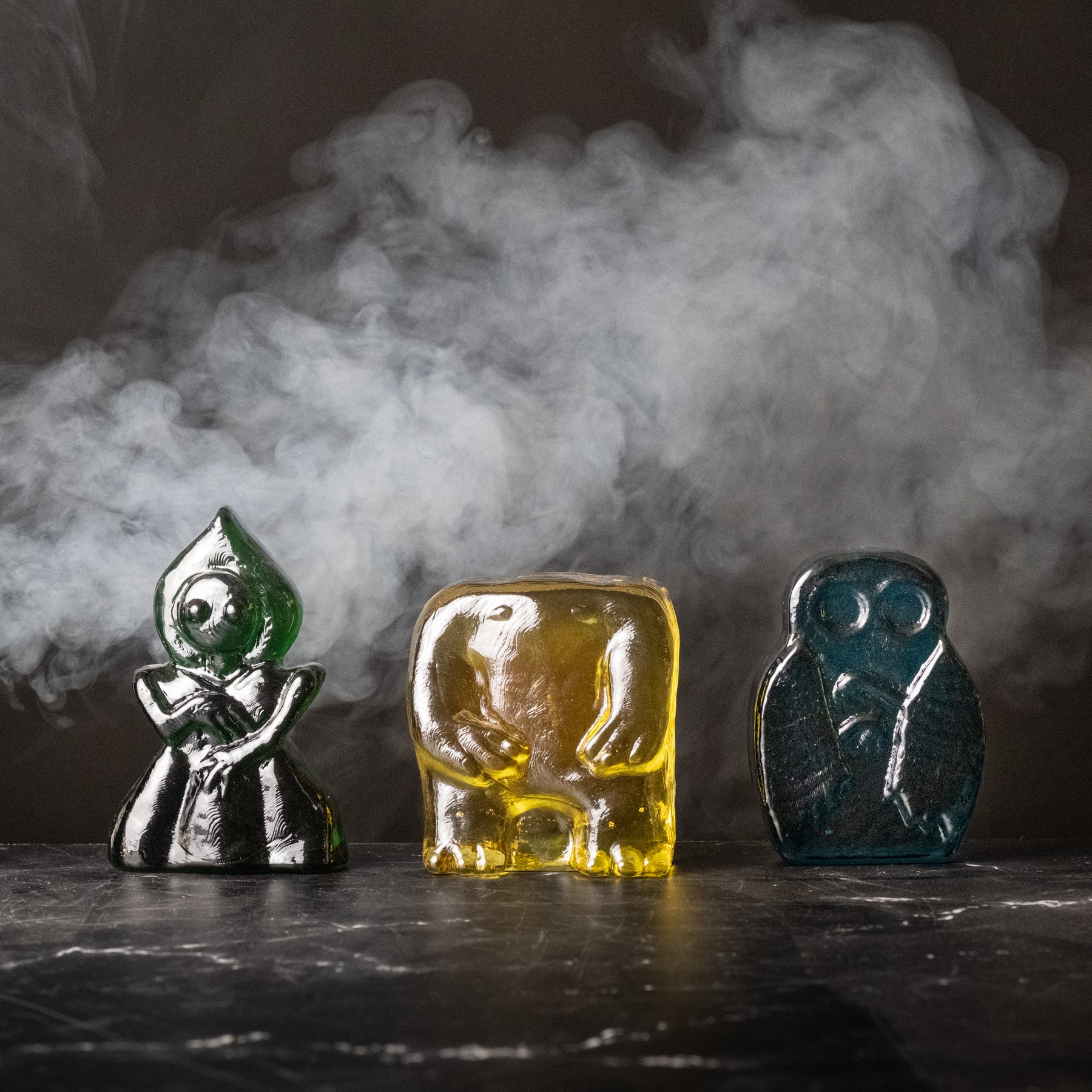 Three glass figurines in green, yellow, and blue on a dark surface with smoke in the background.