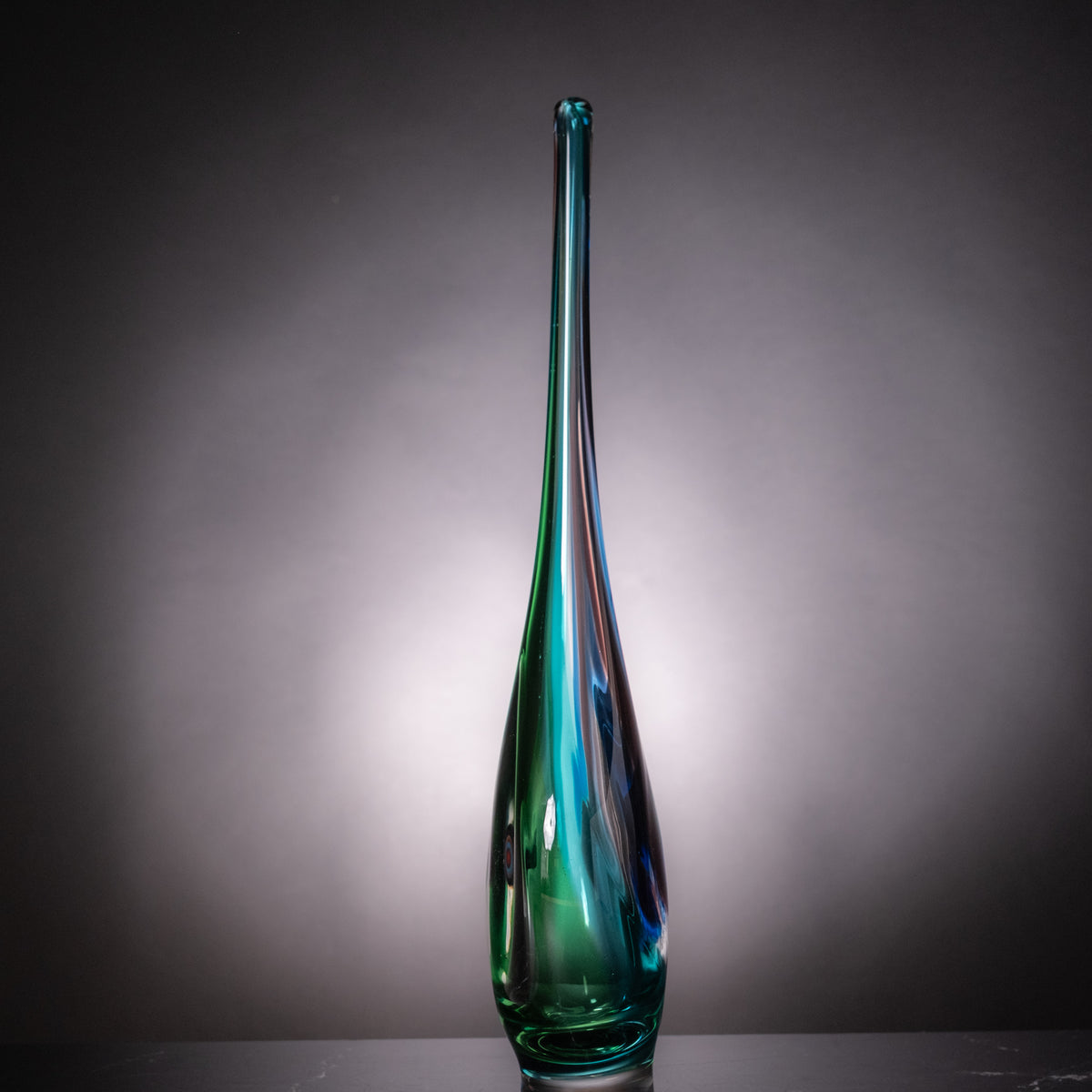 A tall, slender glass vase with a bulbous base, featuring a gradient of green and blue glass with iridescent reflections, set against a dark gray gradient background.
