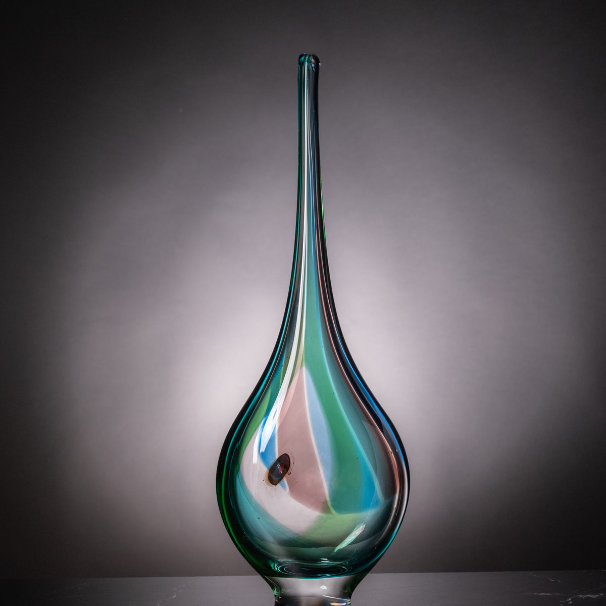 A hand-blown glass vase with a long, slender neck and a bulbous base, featuring iridescent swirls of teal, green, and pink on a dark gray gradient background.