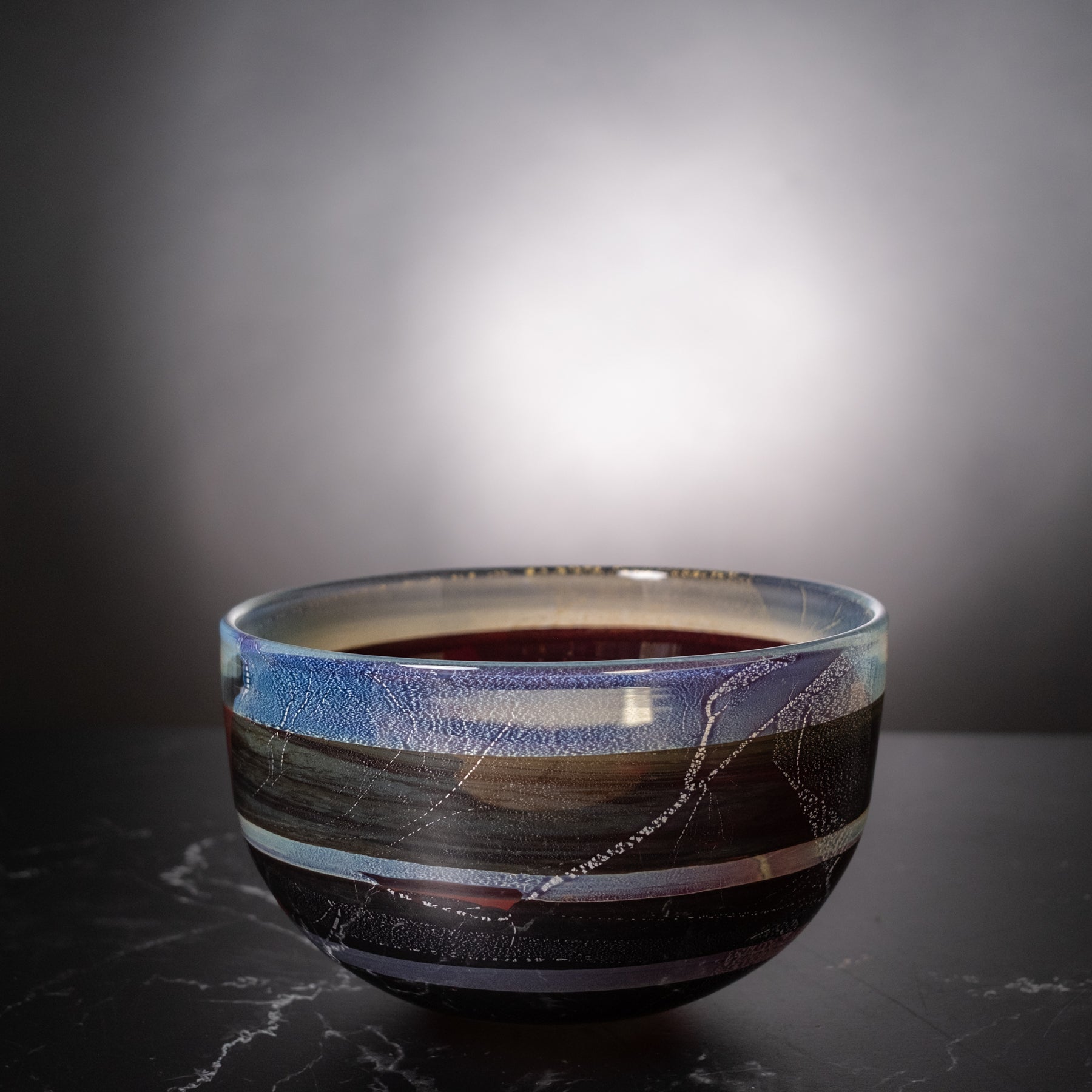 A ceramic bowl with a textured, hand-painted landscape design in shades of blue, brown, black, and white, sitting on a dark marble surface against a dark gray gradient background.