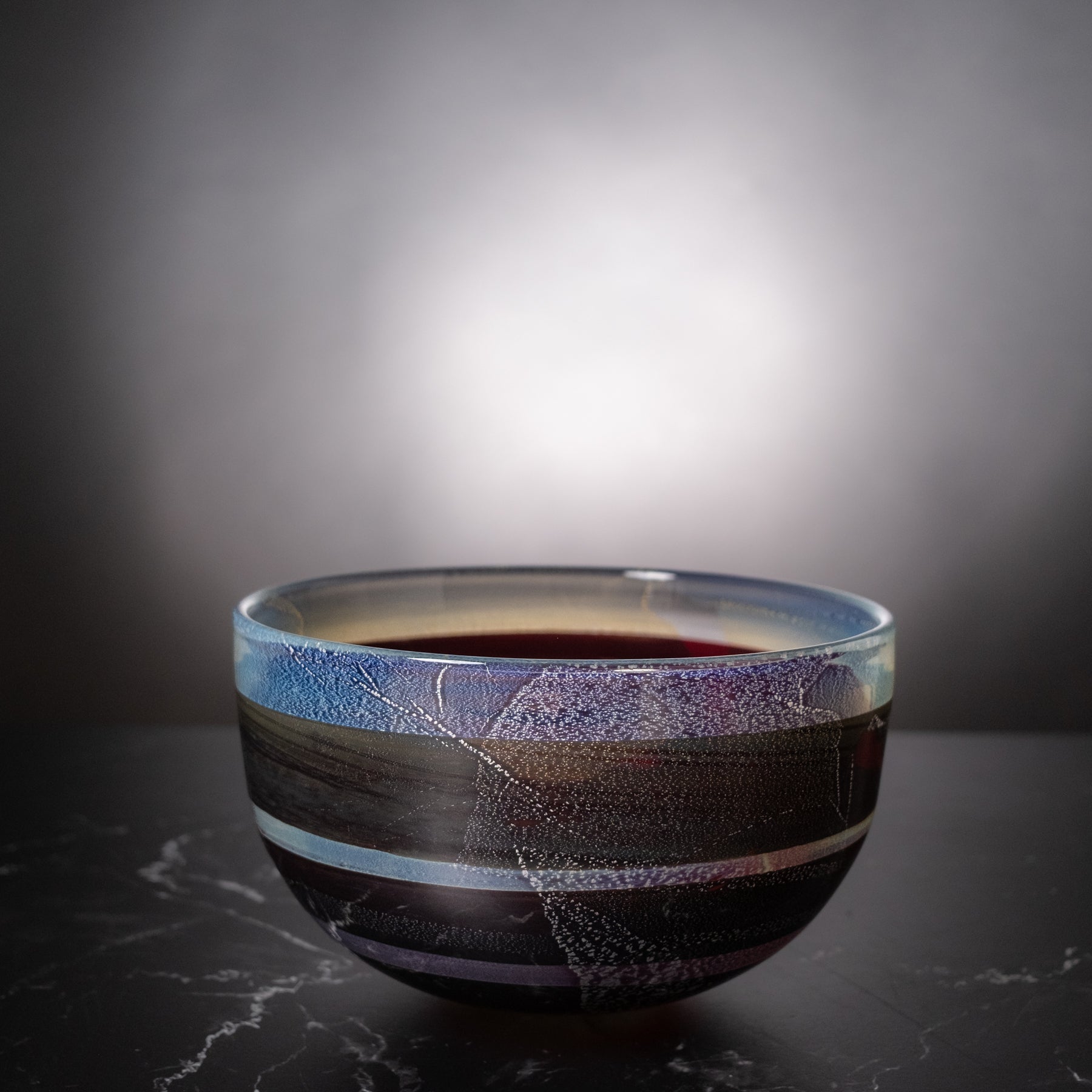 A handmade ceramic bowl with a dark, textured glaze featuring horizontal bands of blue, purple, black, and metallic silver, filled with a dark red beverage, set against a dark gray background.