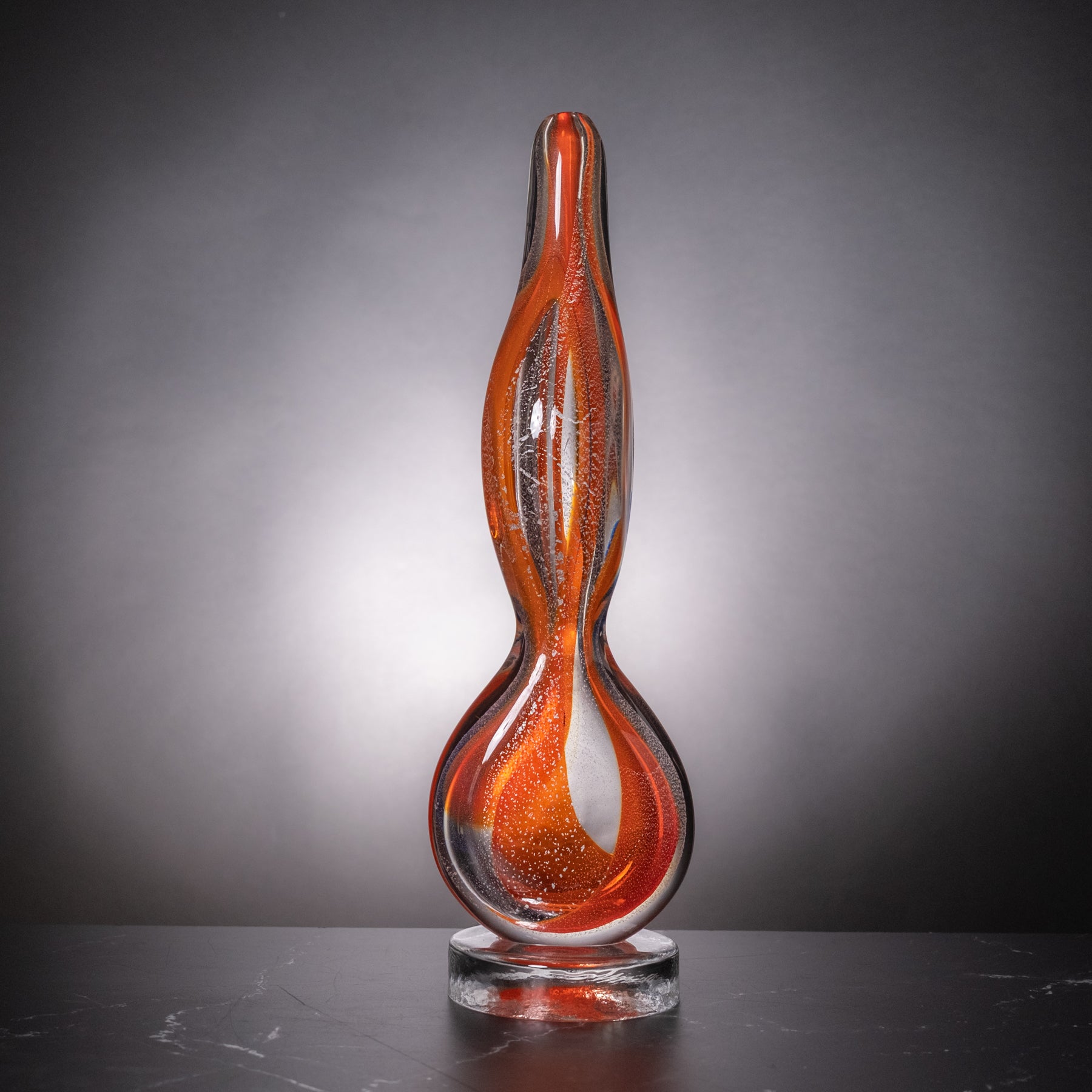 A sculptural, hand-blown glass sculpture with a flame-like silhouette, featuring vibrant orange and red glass with silver and blue accents, displayed on a clear glass base against a dark gray gradient background.
