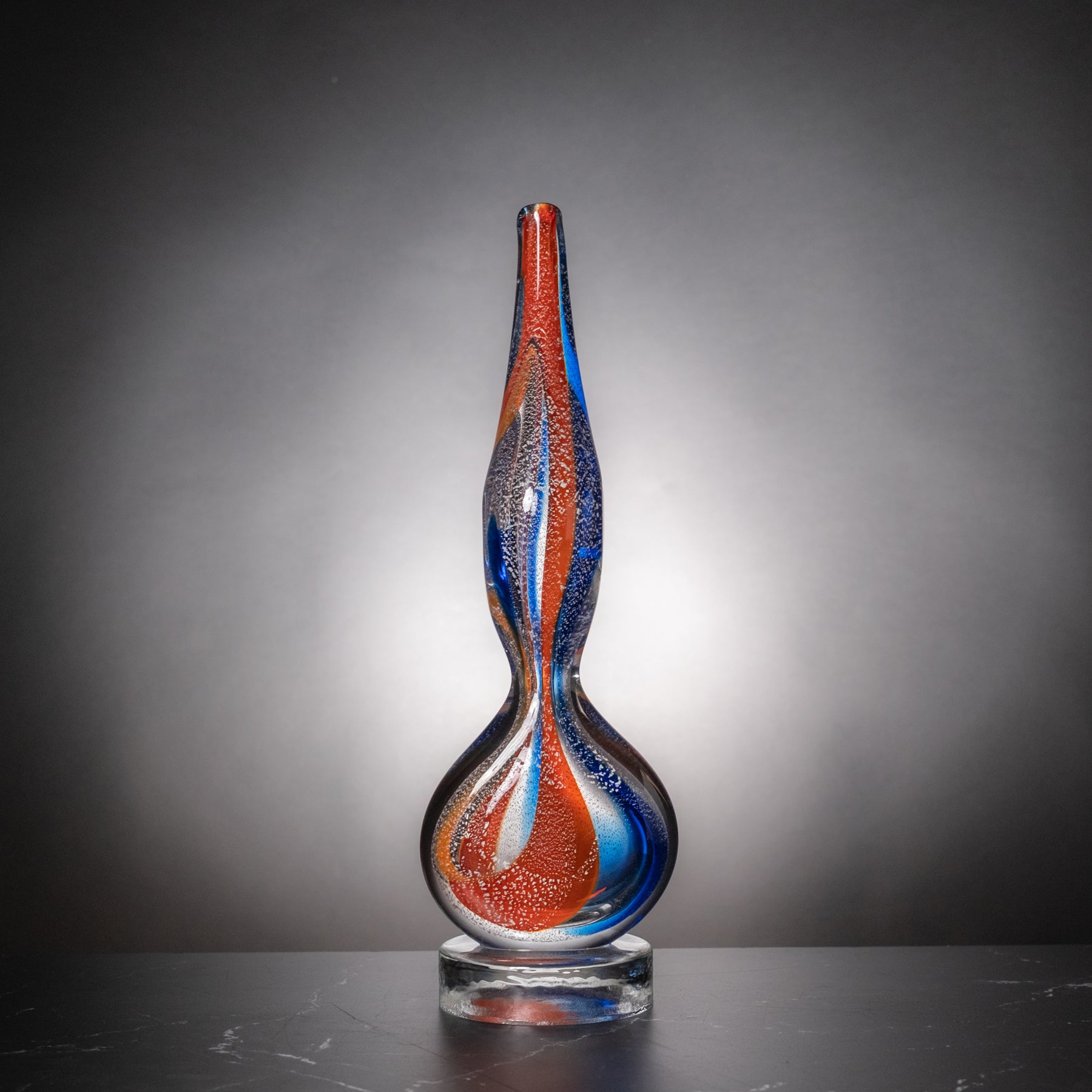 A tall, sculptural glass vase with a narrow neck and bulbous base, featuring swirling red, blue, and clear glass patterns, displayed on a dark surface against a gray gradient background.