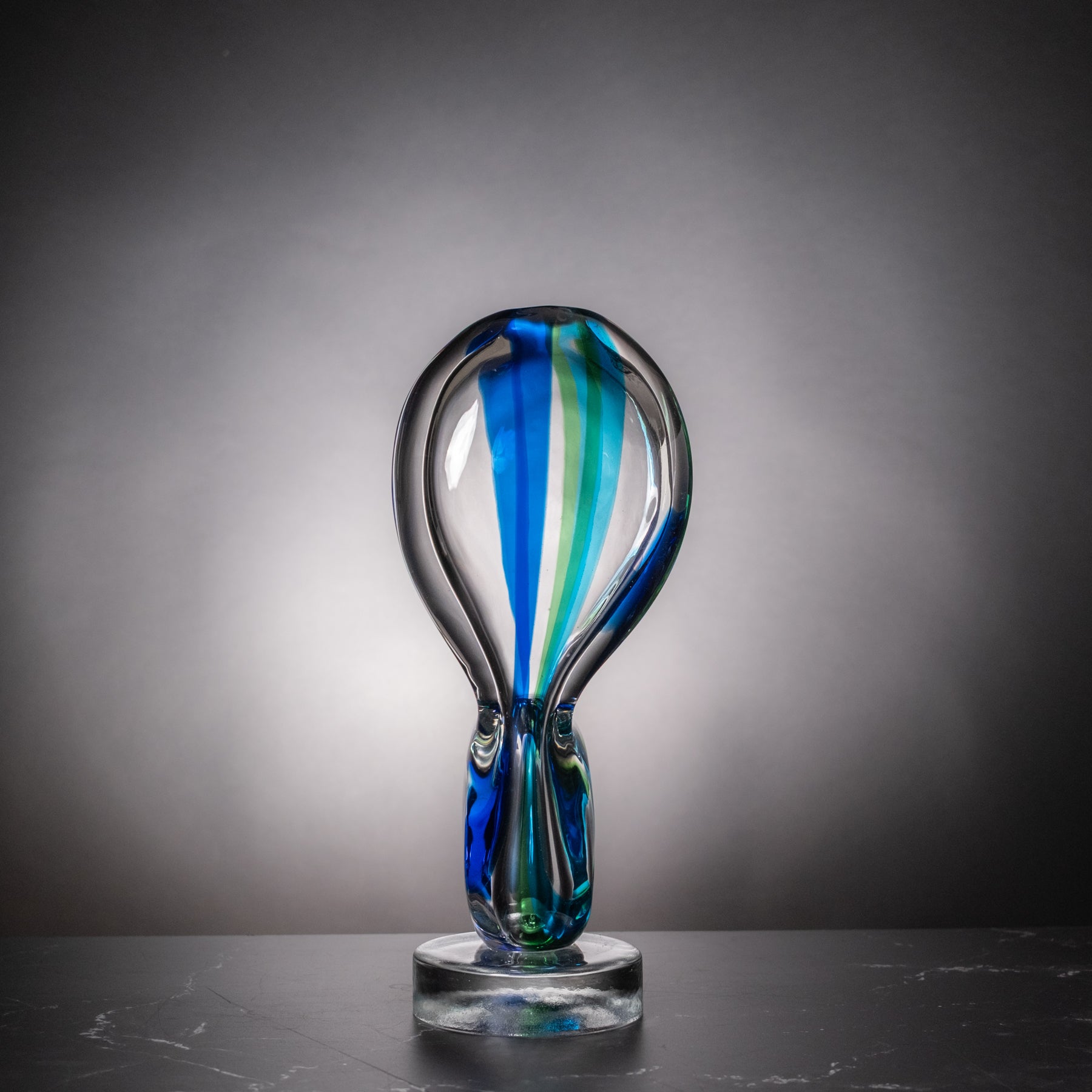 A modern glass sculpture with a teardrop-shaped top featuring vertical blue, green, and white glass swirls, mounted on a clear glass base against a dark gray gradient background.
