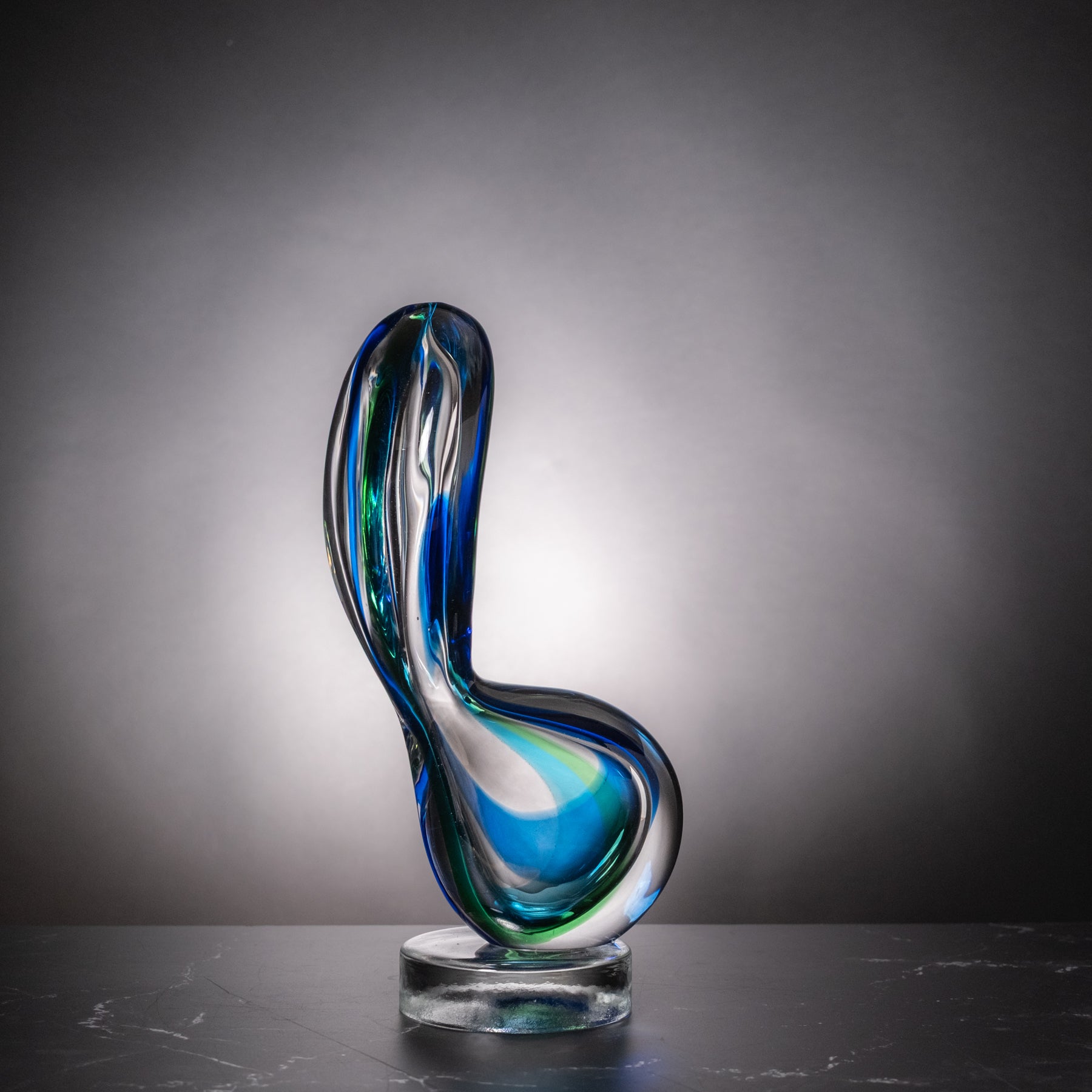 An abstract curved glass sculpture with swirling blue, green, and clear glass layers, displayed on a clear round base against a dark gray gradient background.