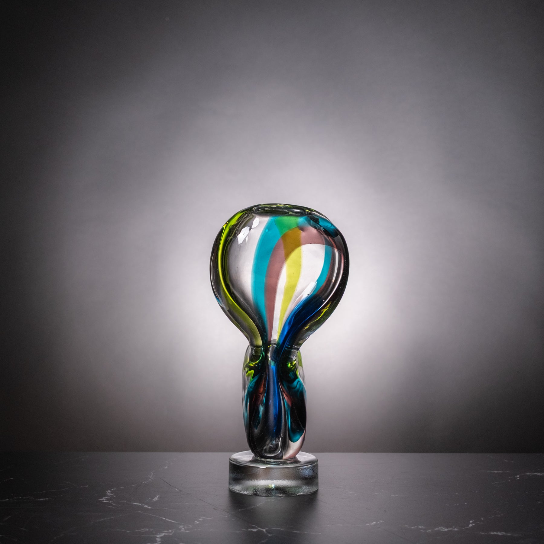 A sculptural glass trophy or award vase with a bulbous top and twisted stem, made from transparent glass with swirling blue, green, yellow, and pink accents, displayed on a dark surface against a gray gradient background.