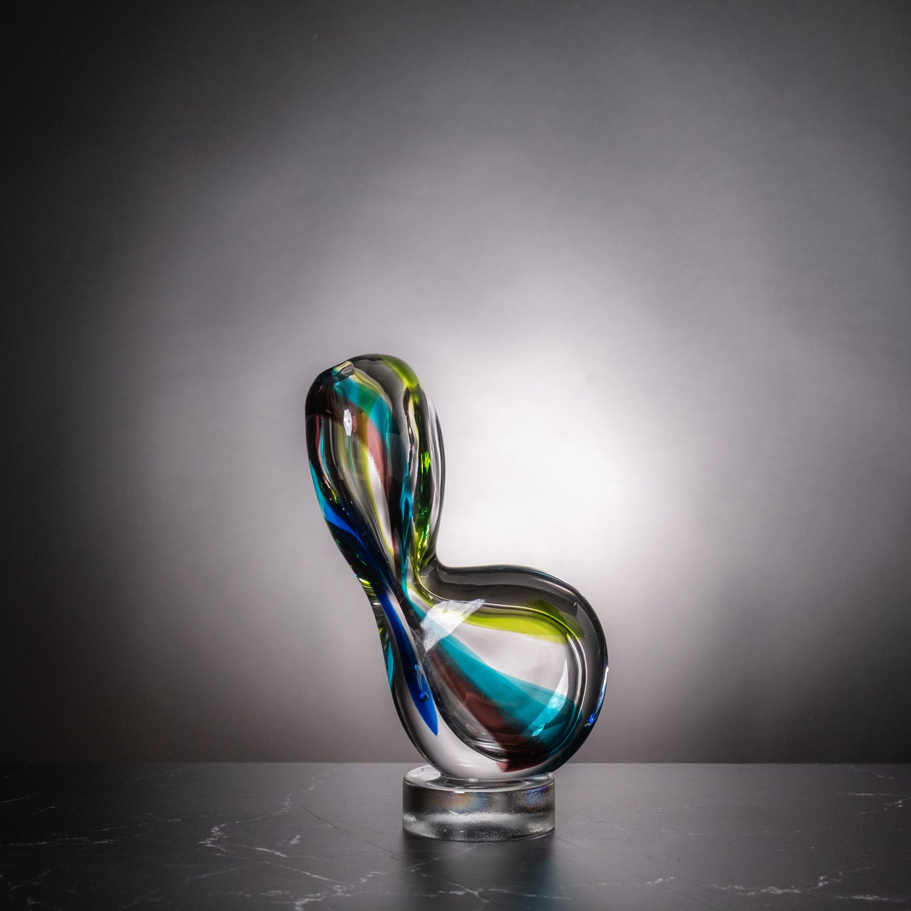 An abstract sculptural glass vase with flowing organic curves, featuring swirls of blue, green, yellow, and purple in transparent glass, displayed on a clear round base against a dark gray gradient background.