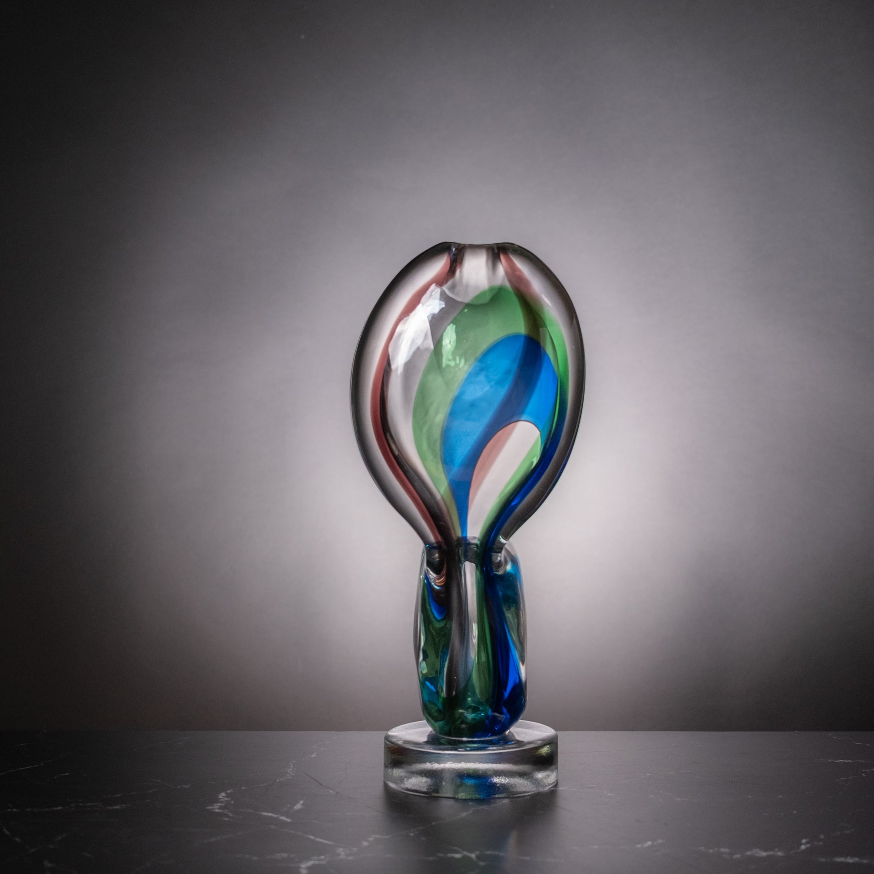 A hand-blown glass sculpture vase with a bulbous top and a cylindrical base, featuring swirls of blue, green, red, and white in clear glass, displayed on a dark surface against a gray gradient background.