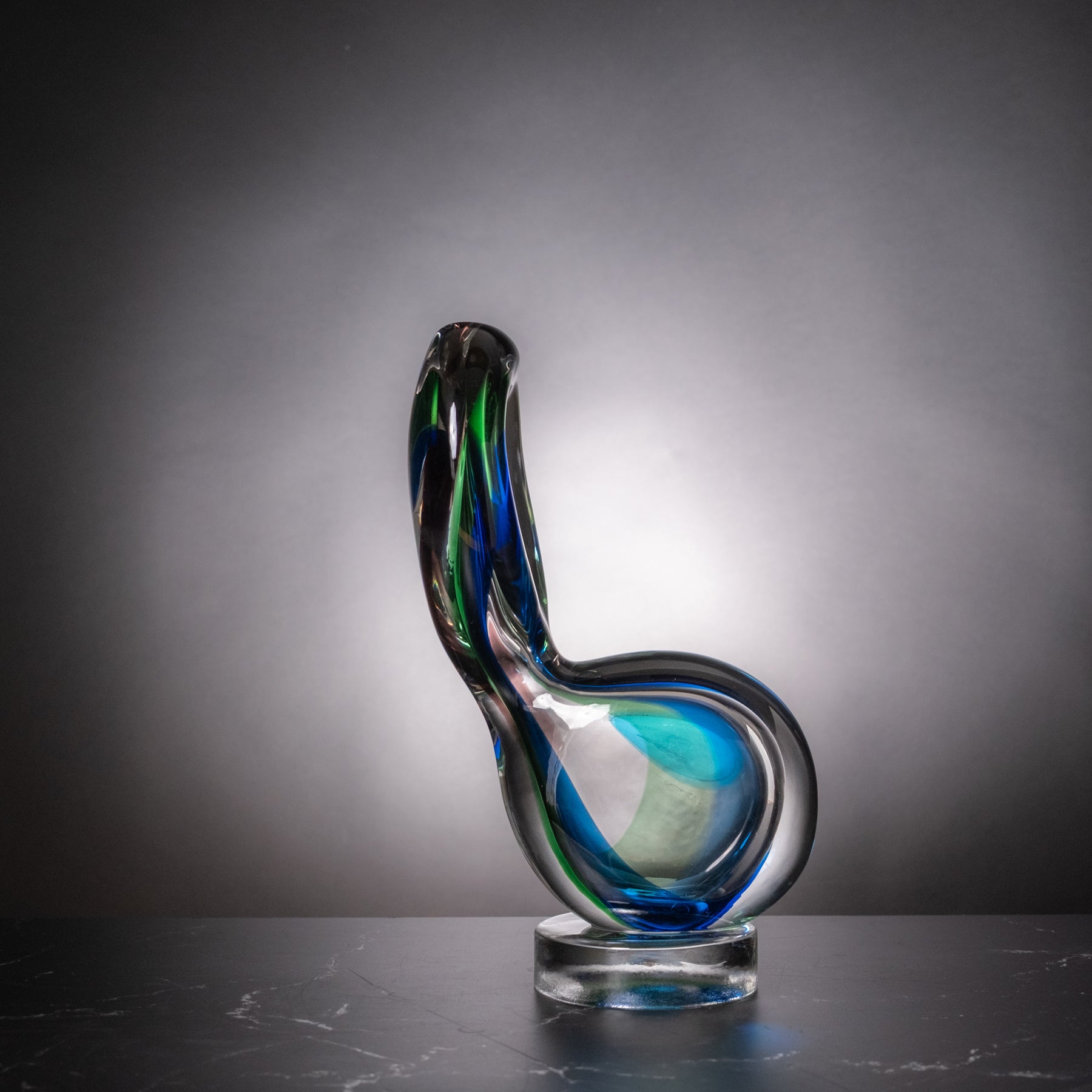 A sculptural glass sculpture with a curved, looped form, featuring swirls of blue, green, and black glass, displayed on a clear round base against a dark gray gradient background.