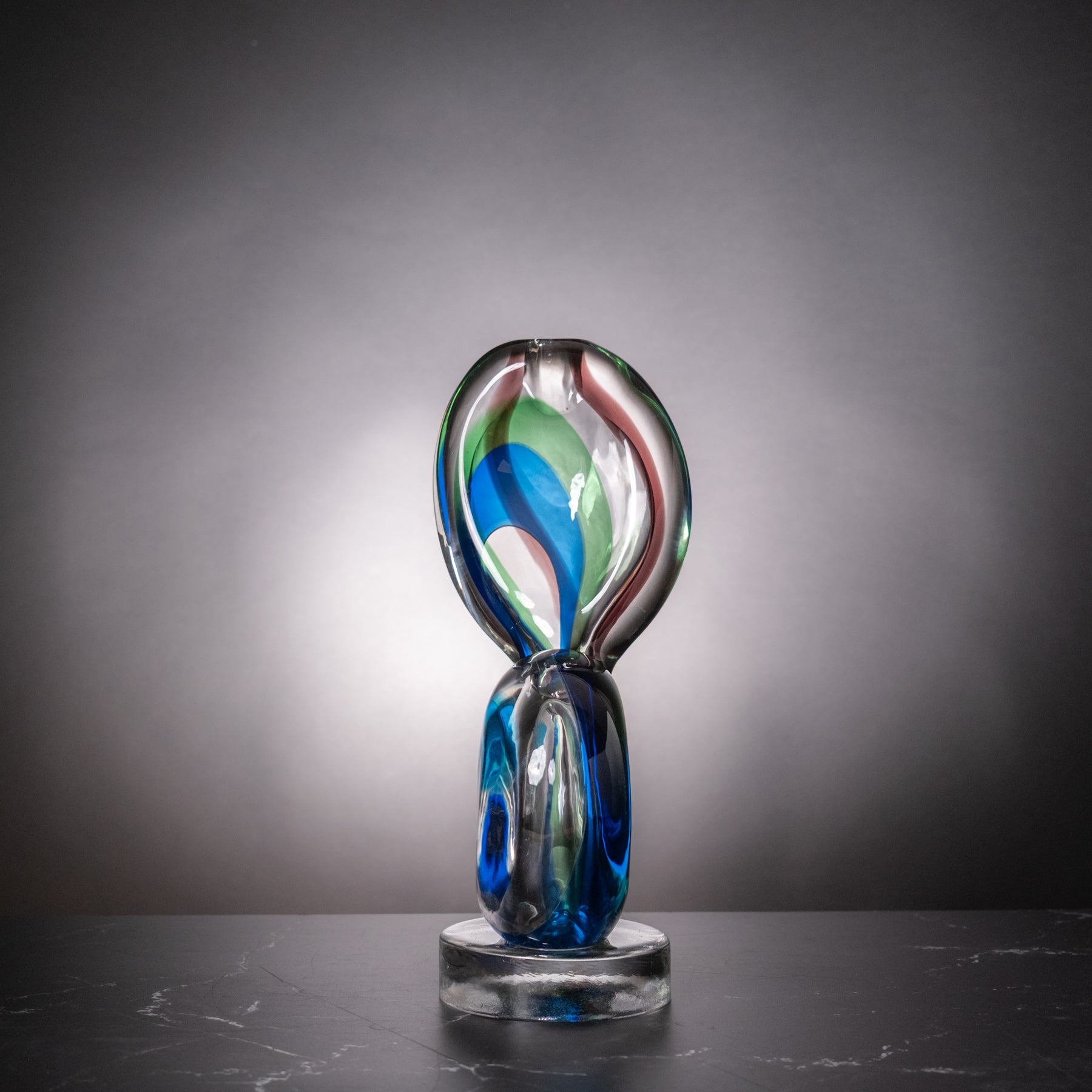 A hand-blown glass sculpture with swirling blue, green, red, and white glass, displayed on a clear glass base against a dark gray gradient background.
