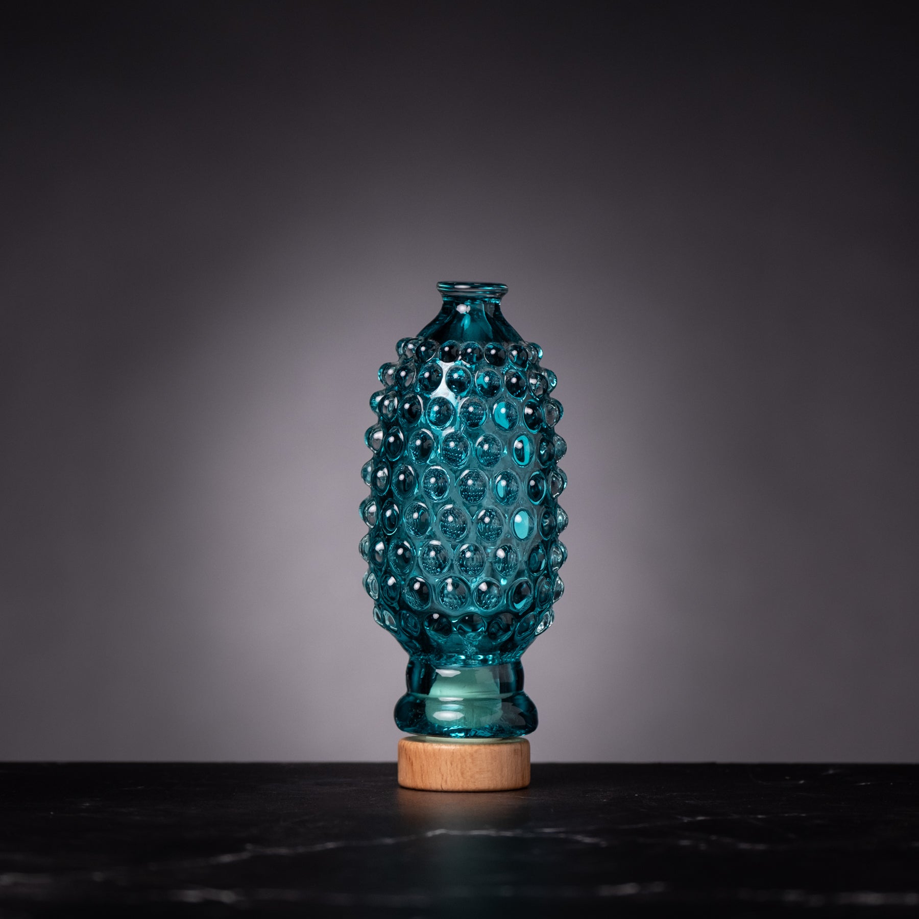 Teal glass vase with wooden base on a dark surface and gray background
