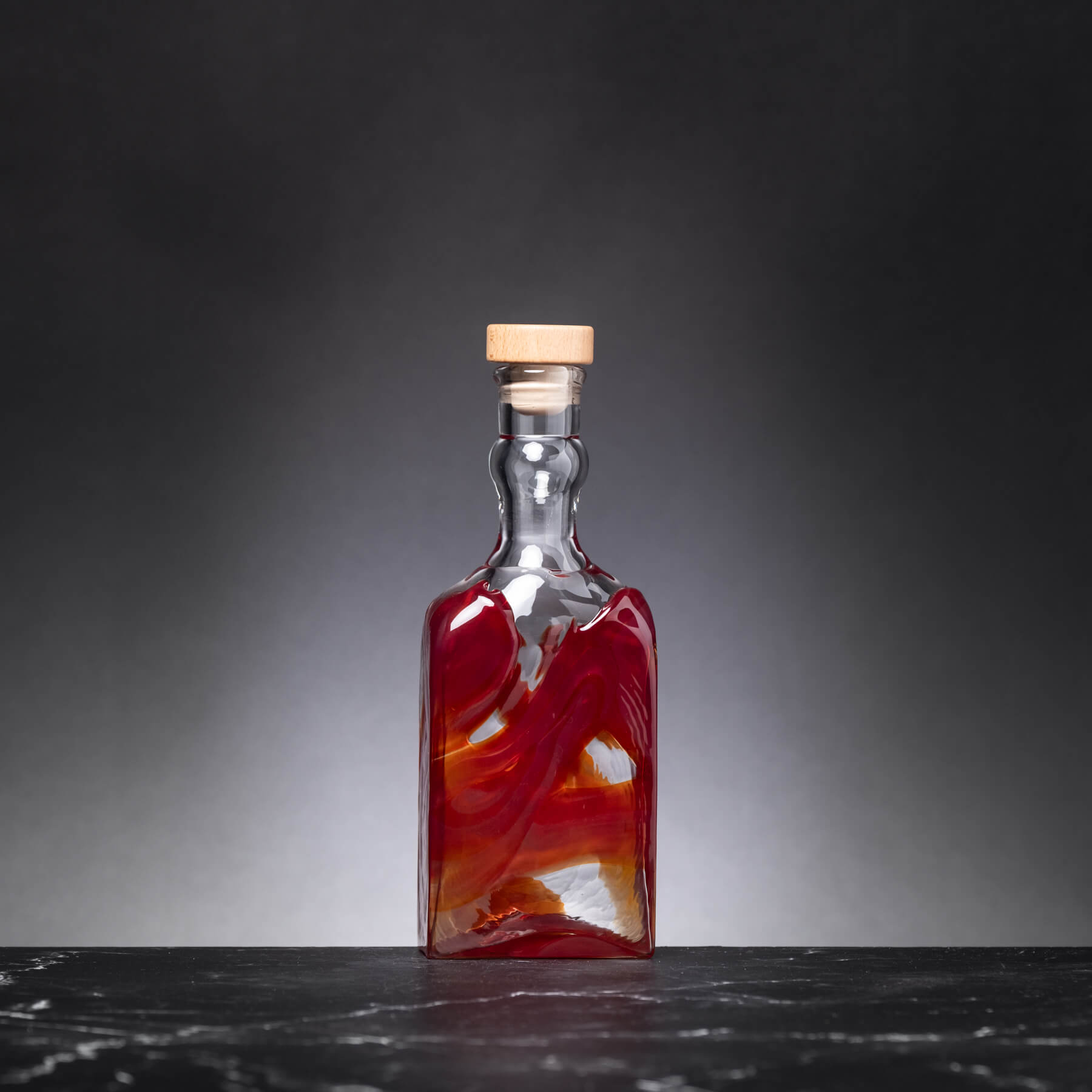 Glass bottle with a red marbled design and wooden cap on a dark surface.