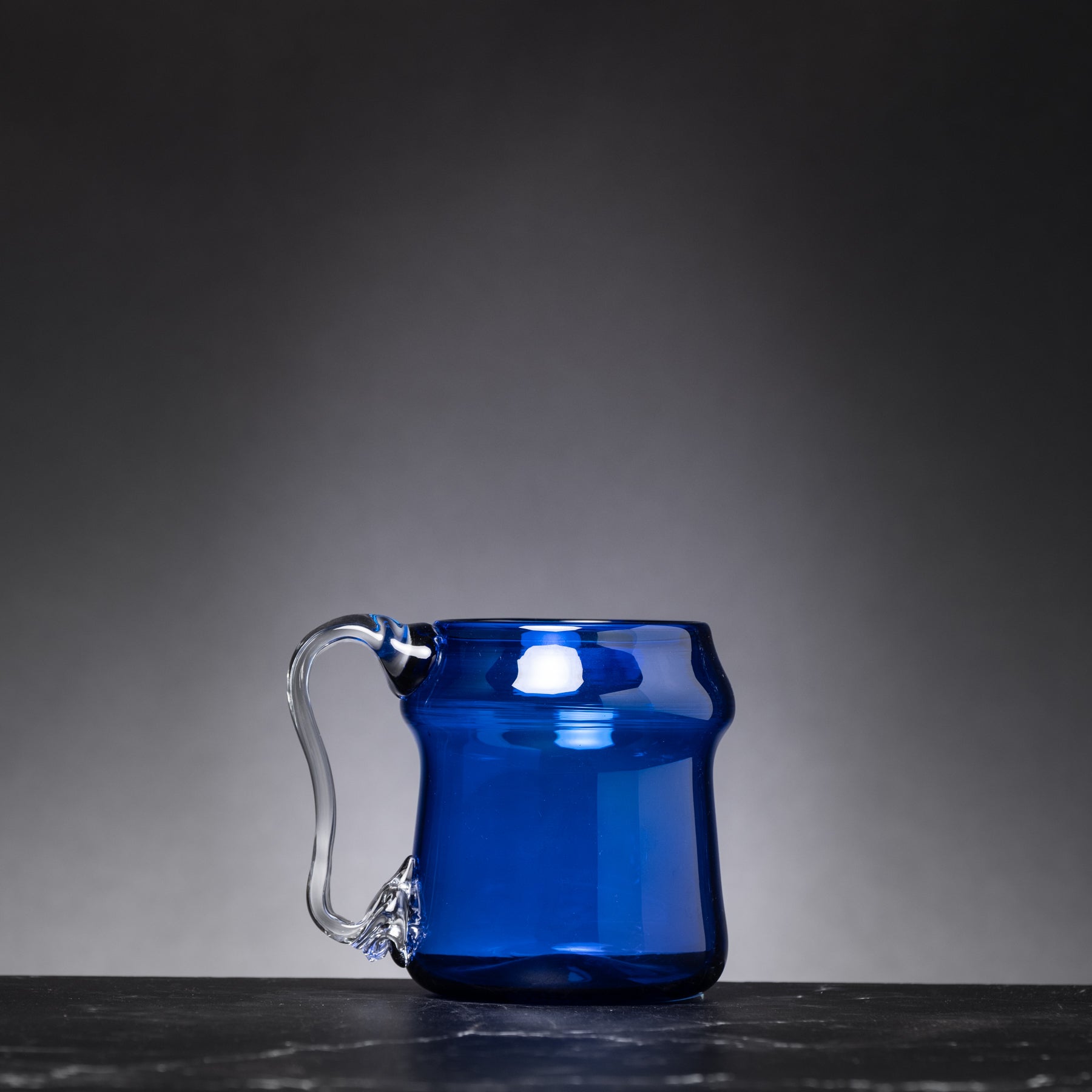 Blue glass mug with a clear handle on a dark surface and gray background