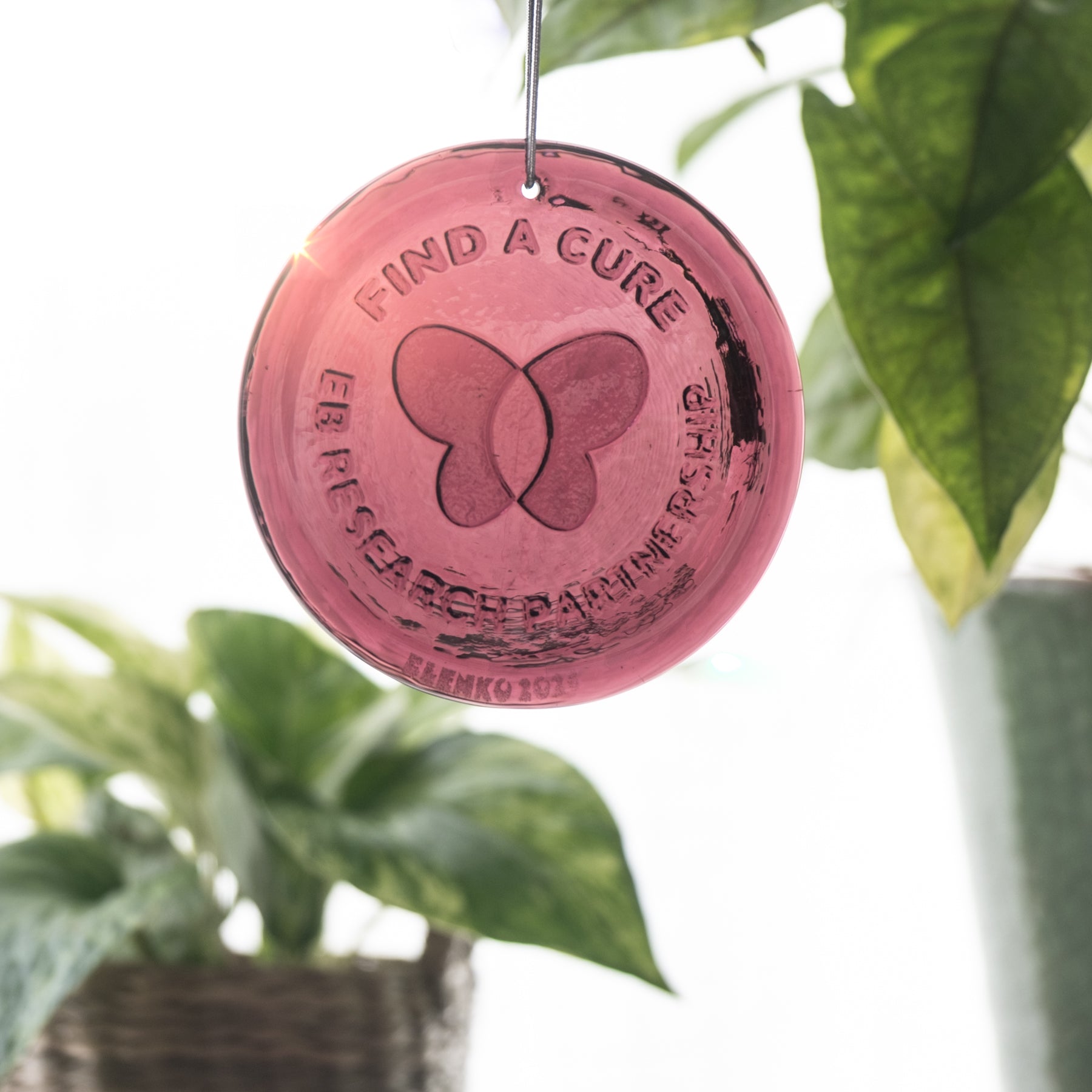 Pink round ornament with butterfly design and 'Find a Cure' text, hanging among green leaves.