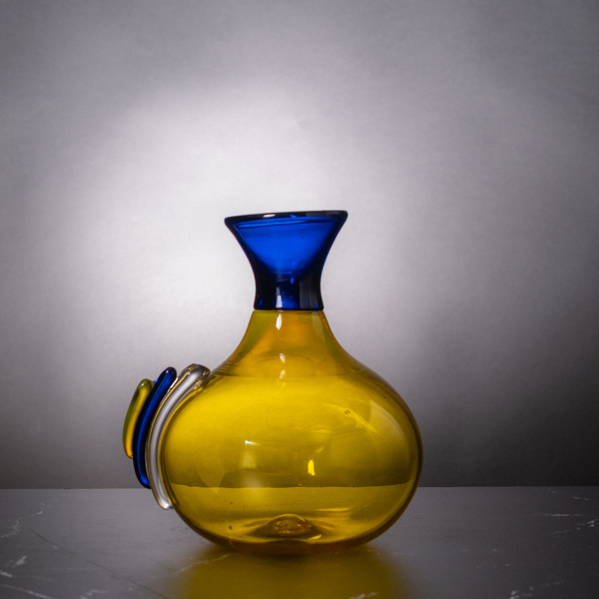 A hand-blown glass vase with a bulbous yellow body, a flared cobalt blue neck, and a matching blue and clear glass side loop, set against a gray gradient background.