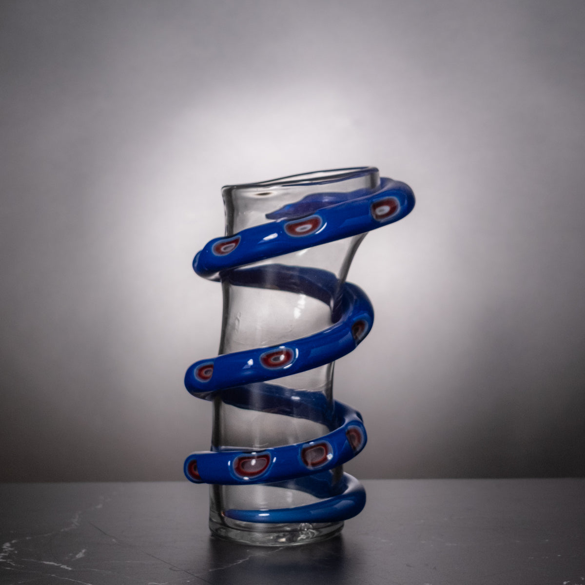 A clear glass vase with a distinctive blue spiral wrap around its body, featuring small red and white accents on the spiral bands, set against a dark gray background.