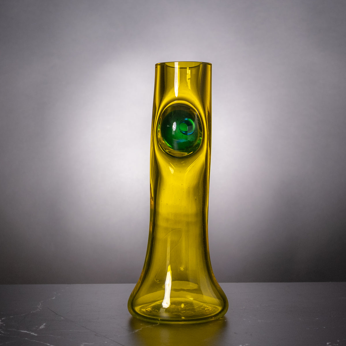 A tall, slender translucent glass vase in yellow-green with a circular green inset near the top, standing on a dark surface against a gray gradient background.