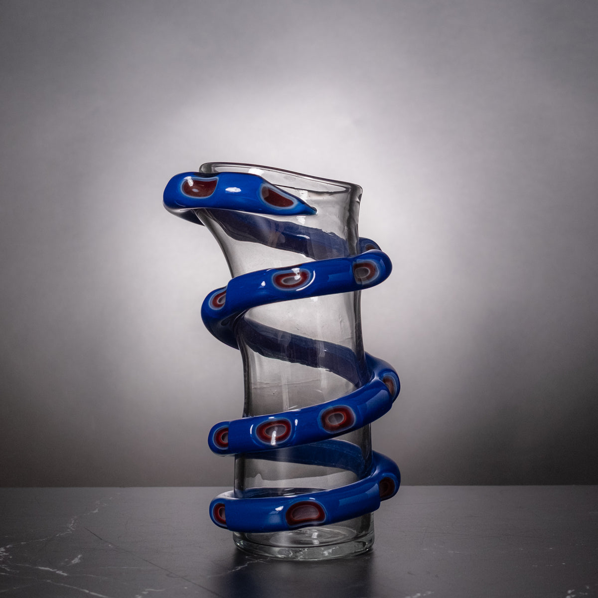 A clear glass vase with a sculptural, twisted spiral design wrapped in glossy blue bands, each featuring small red and white eye-like details, set against a dark gray gradient background.