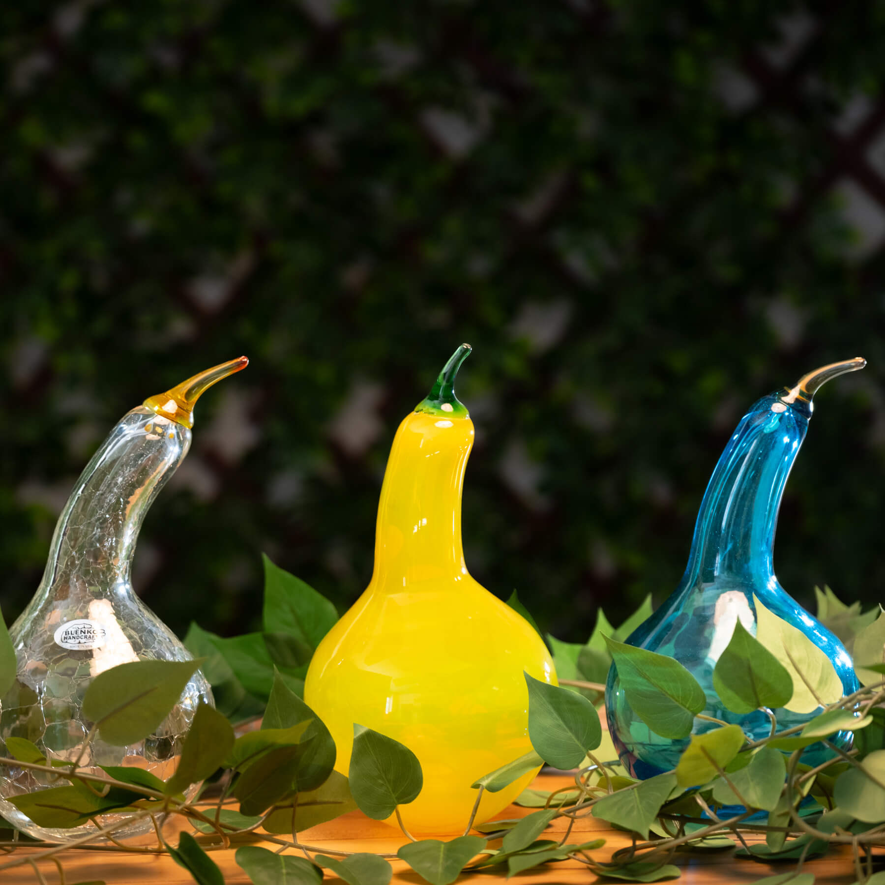 Three glass gourds in yellow, blue, and clear on a leafy background

