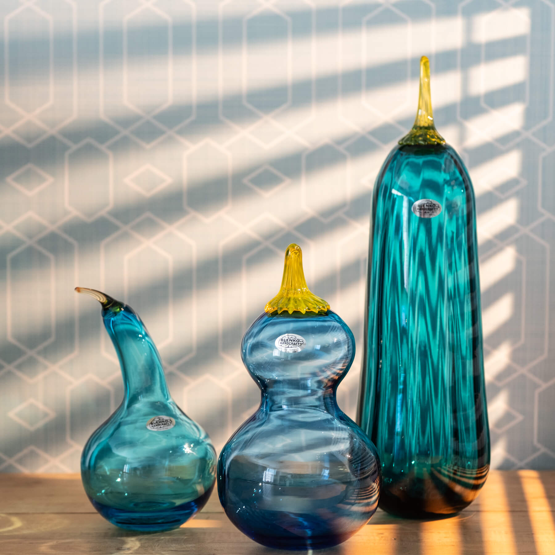 Three blue glass gourds with yellow stems on a wooden surface against a geometric-patterned wall.

