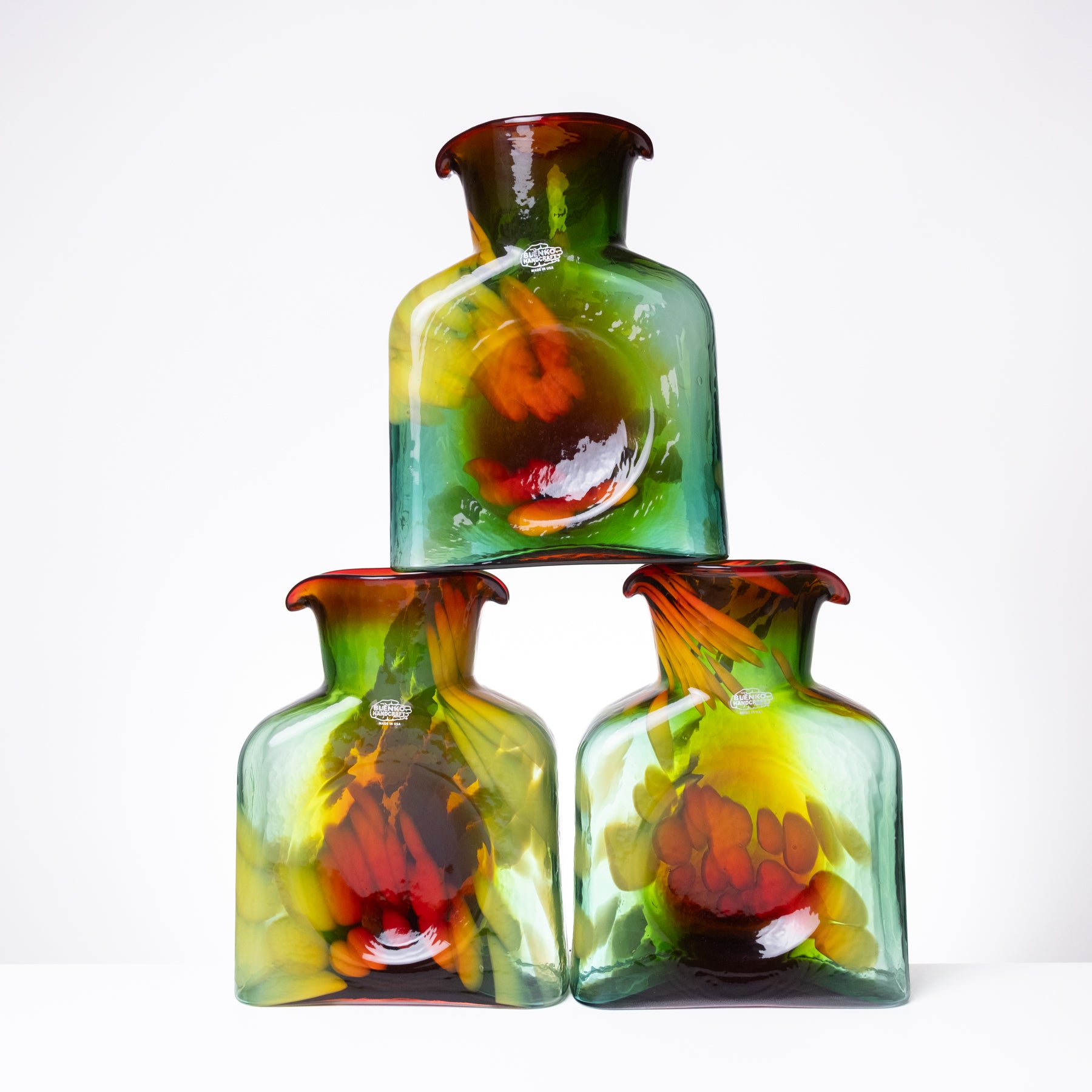 Three hand-blown glass bottles stacked in a pyramid arrangement on a white background. The bottles feature a translucent green body with vibrant, swirling internal patterns of yellow, orange, and red, and a dark amber-brown rim.
