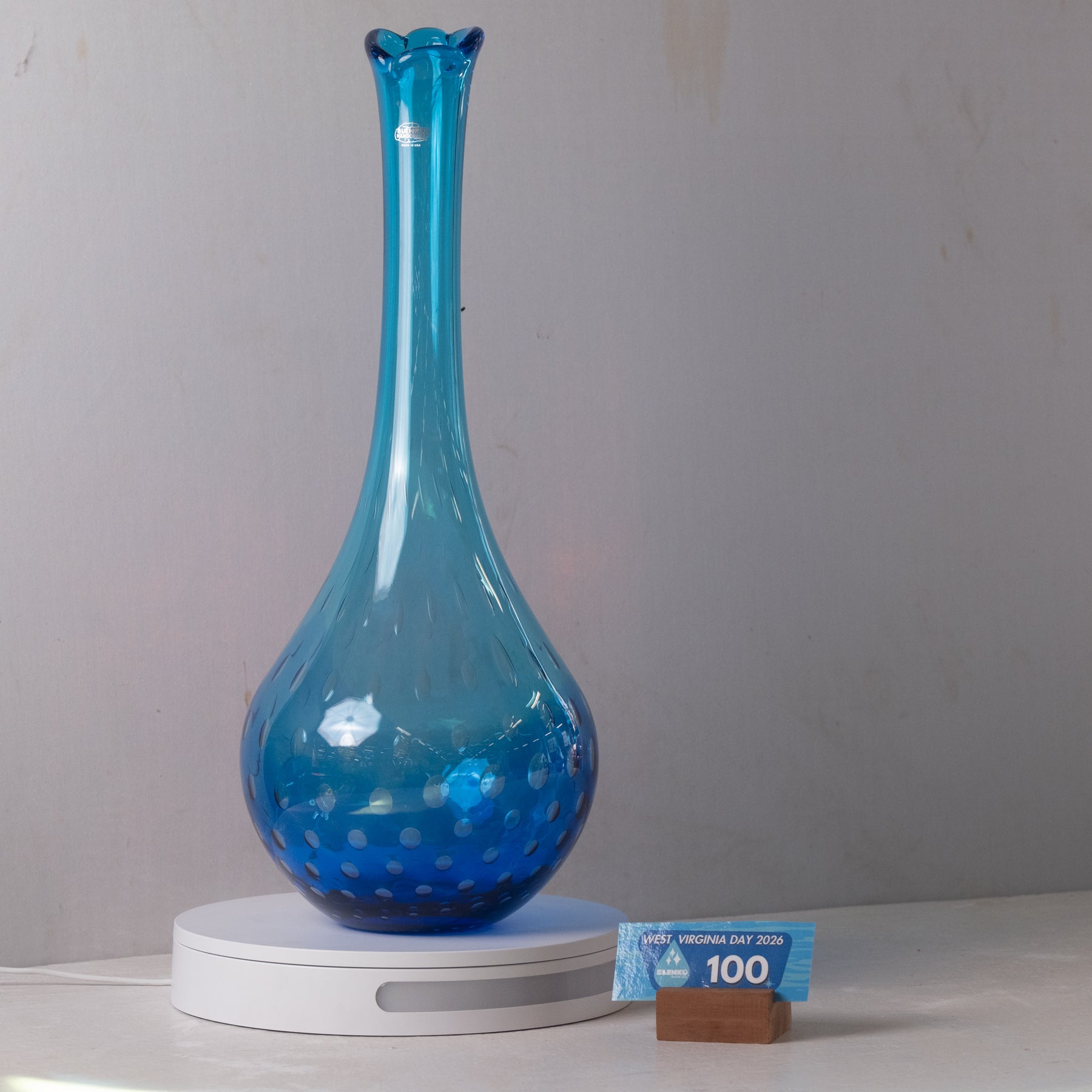 A translucent cobalt blue glass vase with a flared neck and bubble-patterned base, displayed on a white circular scale against a light gray wall. A small blue and brown price tag stands beside the base.