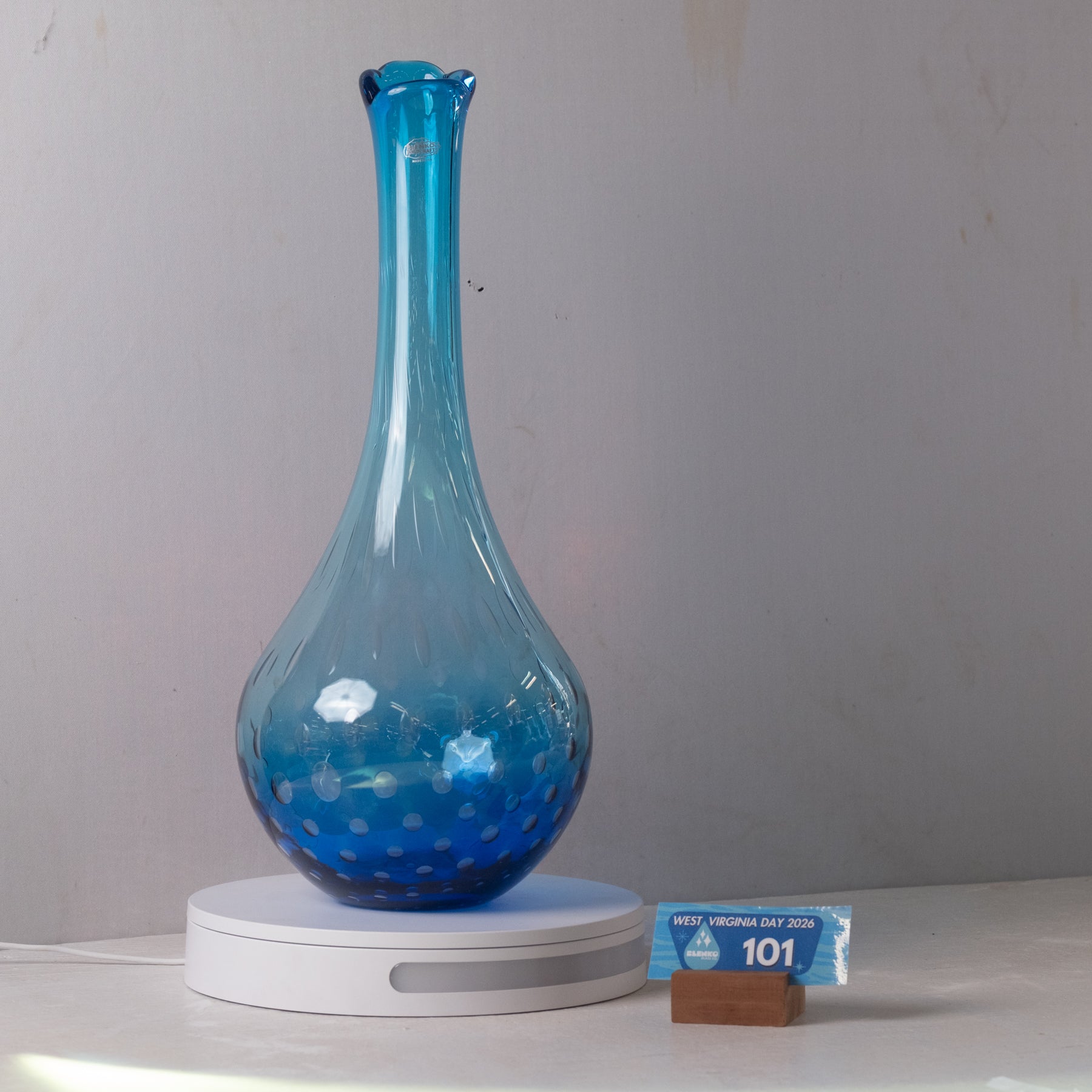 A translucent blue glass vase with a flared neck and dotted pattern near the base, displayed on a white circular charging base against a light gray wall.