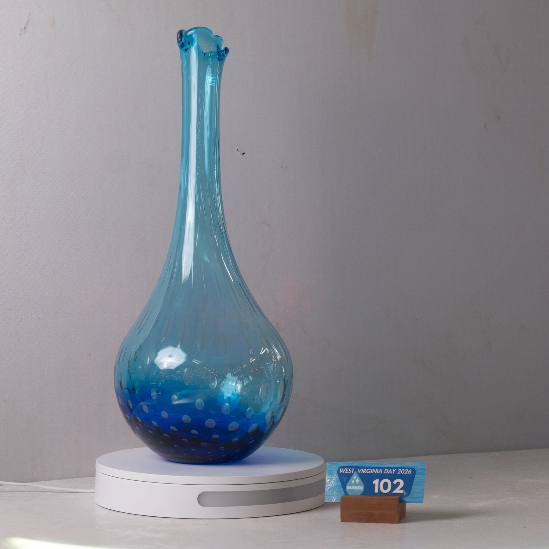 A translucent blue glass vase with a flared neck and speckled pattern near the base, displayed on a white circular charging base against a plain light gray wall. A small blue price card on a wooden block sits to the right.