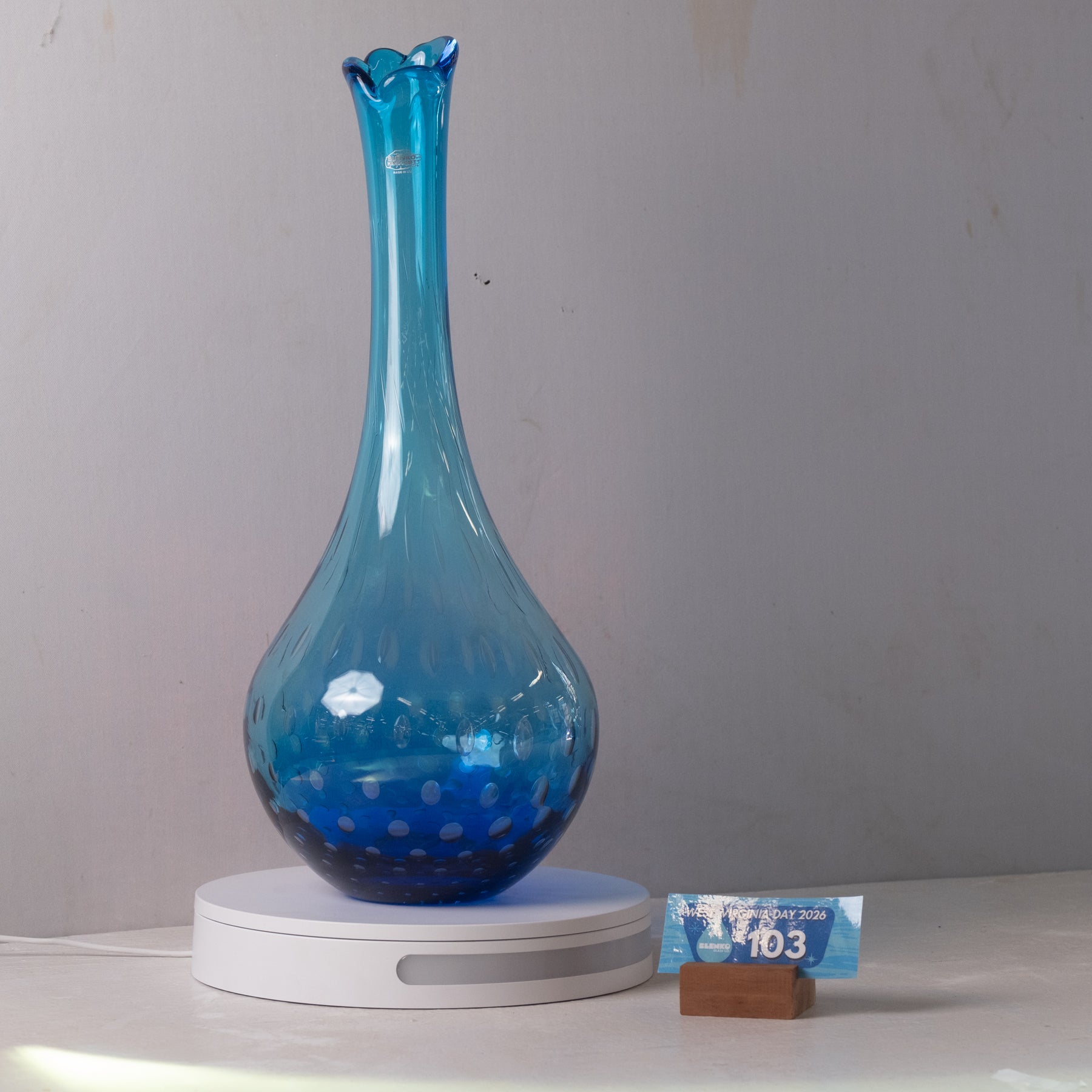 A translucent blue glass vase with a flared neck and dotted pattern near the base, displayed on a white circular charging base against a plain light gray wall.