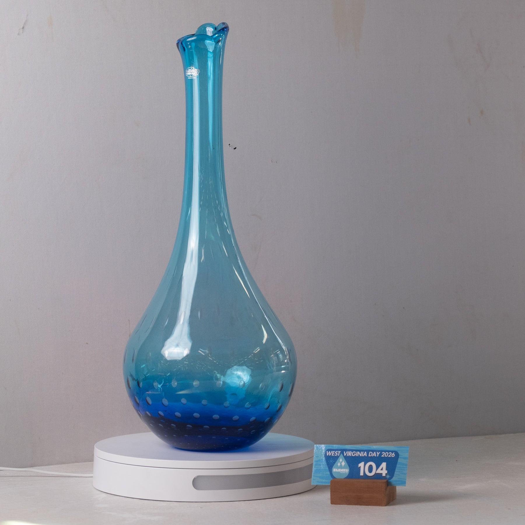 A translucent blue glass decanter with a long neck and rounded base, displayed on a white circular charging base against a plain light gray wall.