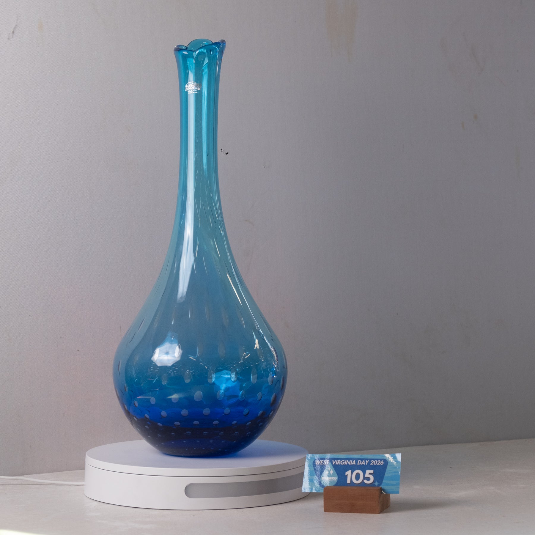 A translucent blue glass vase with a bulbous base and narrow flared neck, displayed on a white circular charging or heating base against a plain light gray wall. A small blue price tag on a wooden block reads $10.