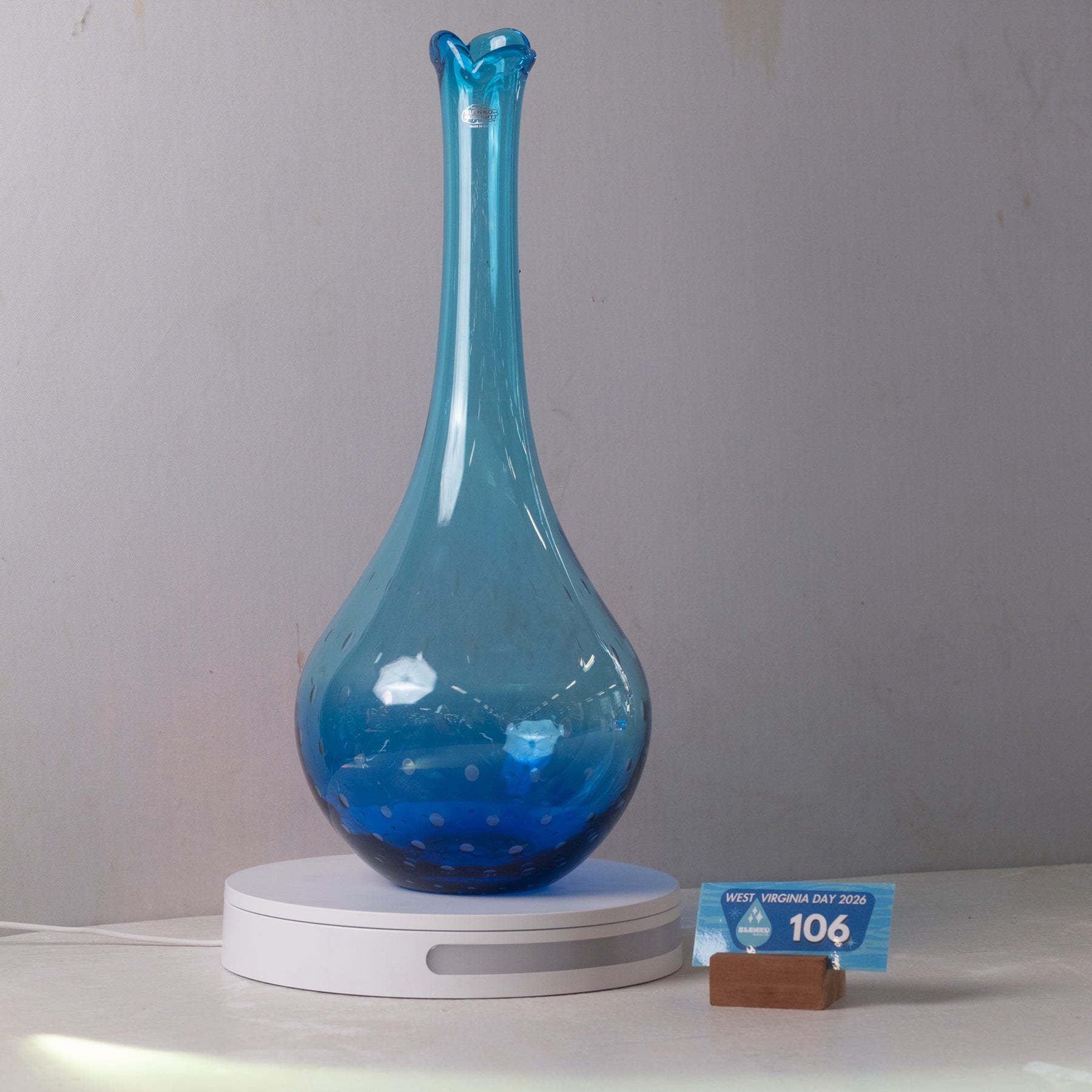 A translucent blue glass vase with a flared neck sits on a white circular charging base. A small blue price tag card is placed beside the base on a white surface against a plain light gray wall.