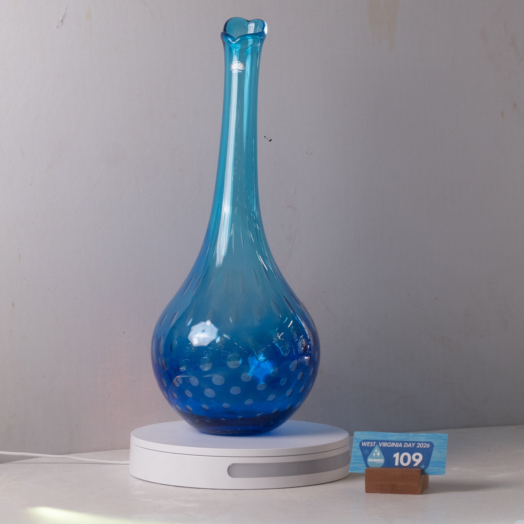 A translucent blue glass vase with a bulbous base and narrow neck, displayed on a white circular light box. A small blue price tag card is visible to the right against a plain light gray wall.