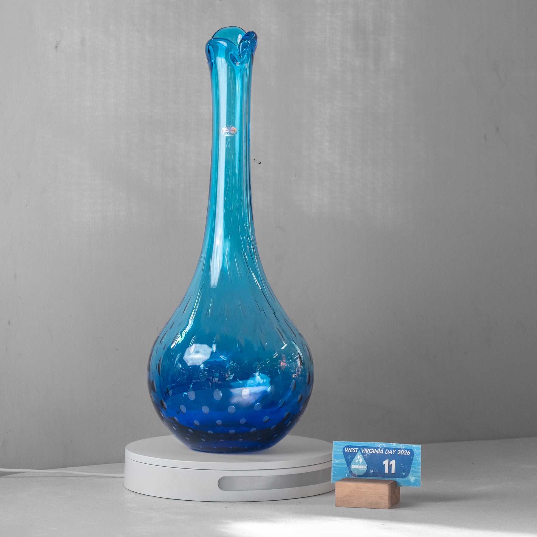 A translucent blue glass decanter with a bulbous base and narrow neck, displayed on a white circular charging base against a gray wall. A small blue card on a wooden block sits to the right for scale.