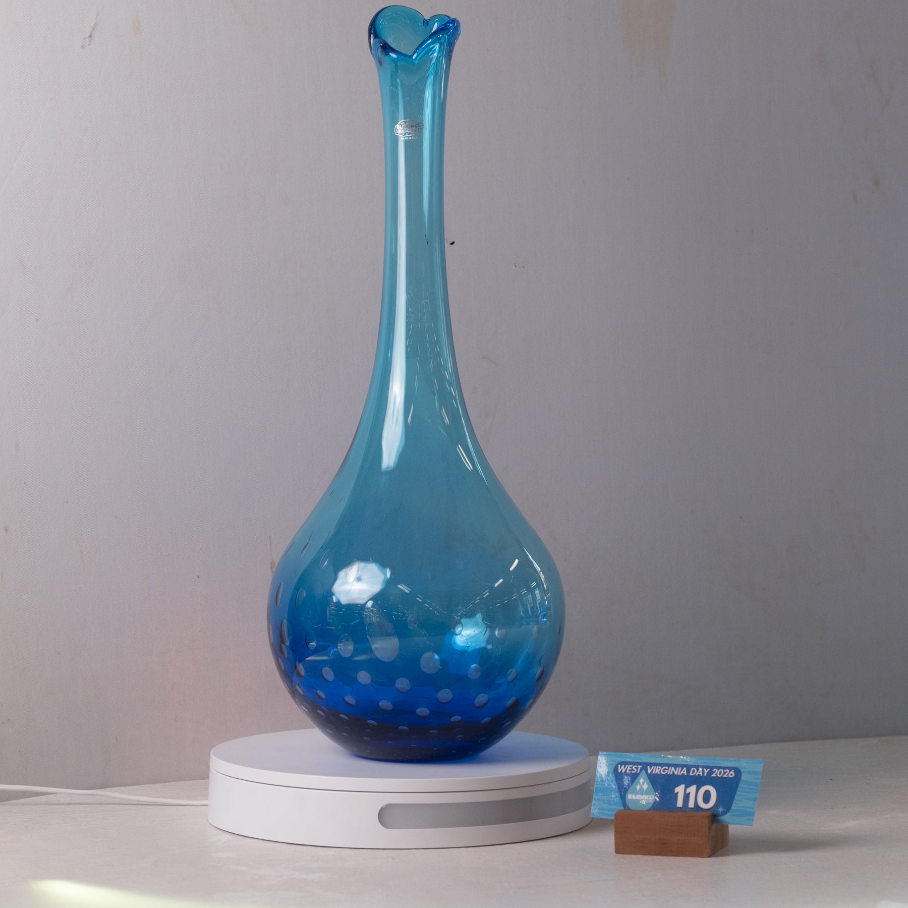 A blue glass vase with a flared neck and small white speckles near the base, displayed on a white circular base against a light gray wall. A small blue price tag and brown block are visible beside the base.