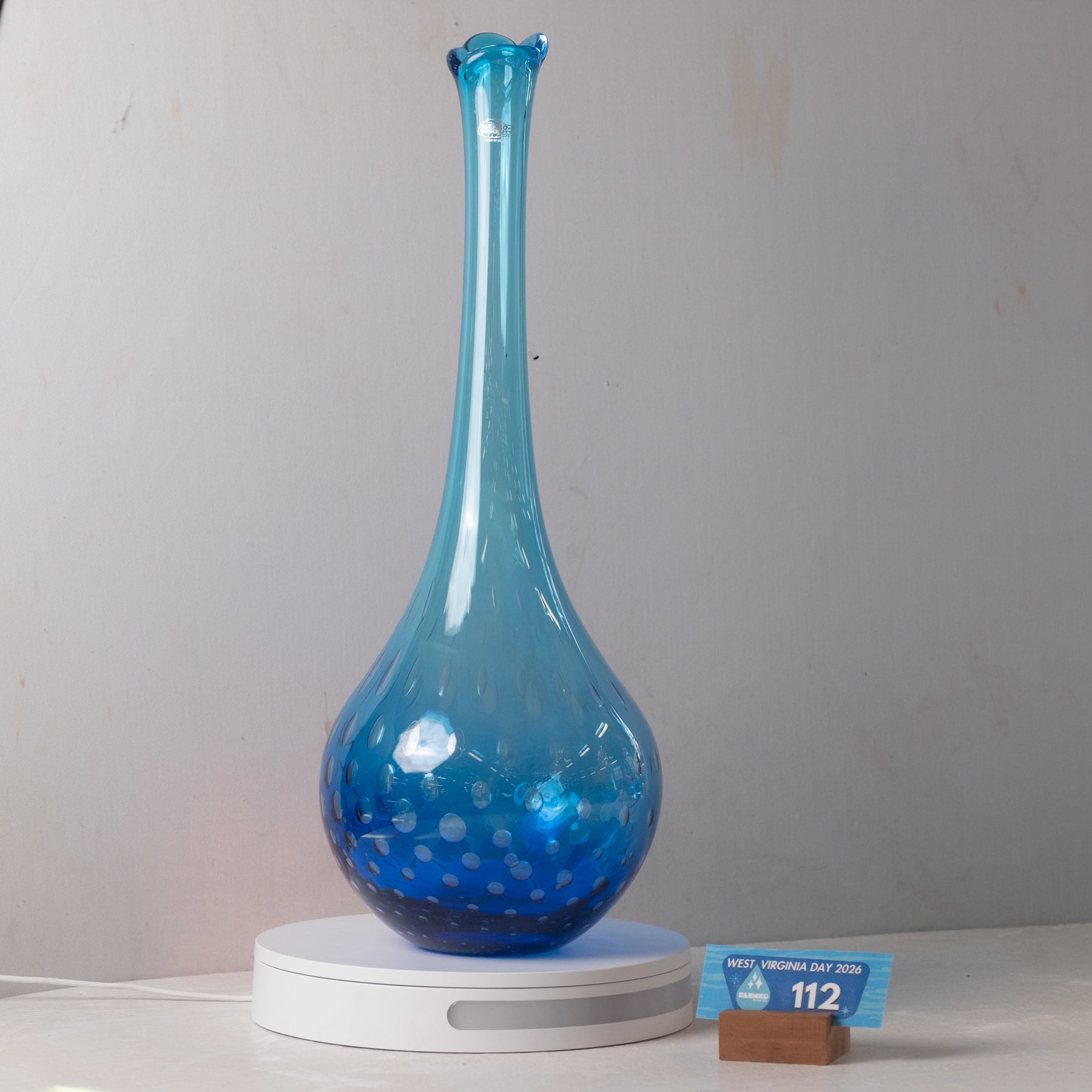 A blue glass vase with a flared neck and speckled pattern, displayed on a white circular light box with a small blue price tag in front, set against a plain gray wall.