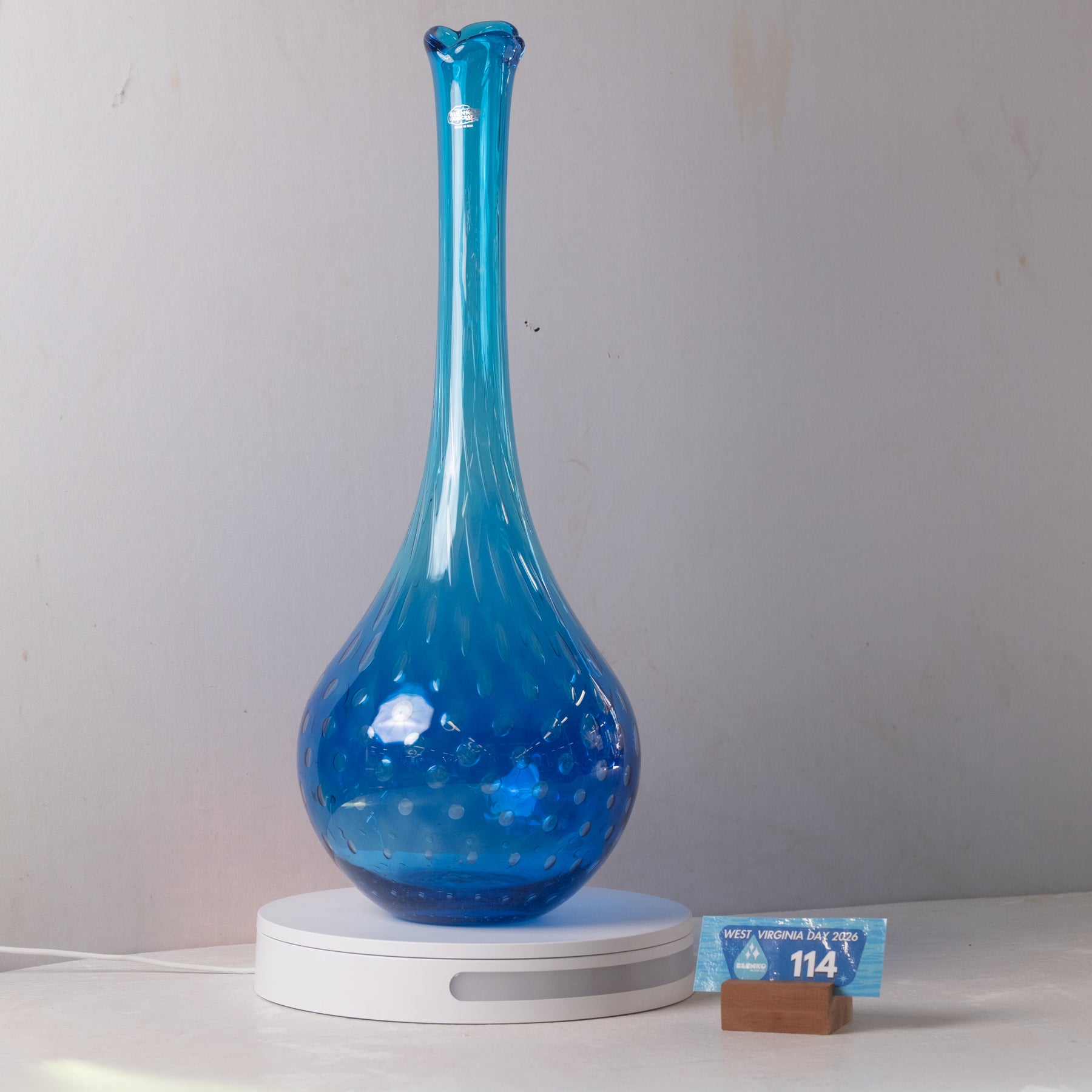 A translucent blue glass vase with a flared neck and dotted pattern near the base, displayed on a white circular light box. A small wrapped item and a brown block are placed beside it on a white surface against a plain light gray wall.