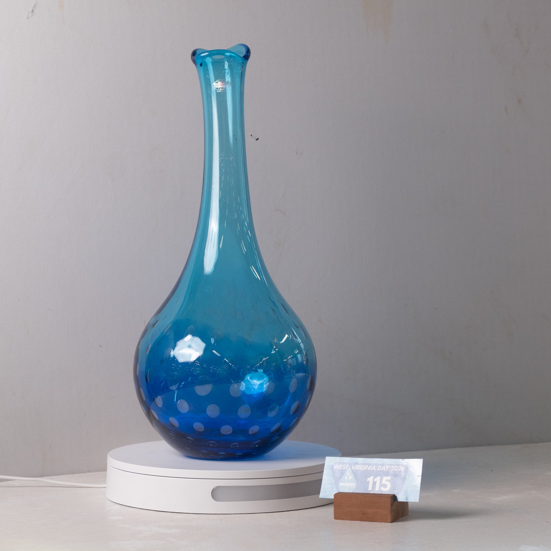 A translucent blue glass vase with a flared neck and dotted pattern near the base, displayed on a white circular light box. A small wrapped item and a brown block are placed beside it on a white surface against a plain light gray wall.