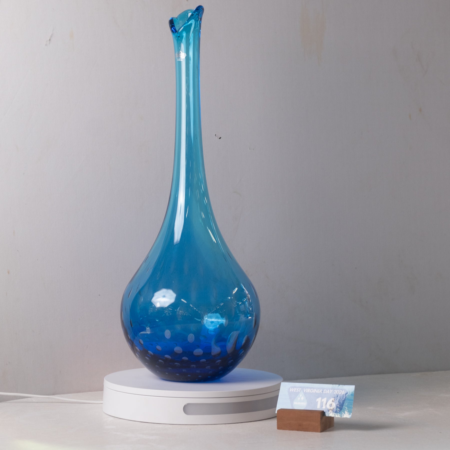 A blue glass vase with a flared neck and spotted pattern near the base, displayed on a white circular stand against a plain light gray wall.
